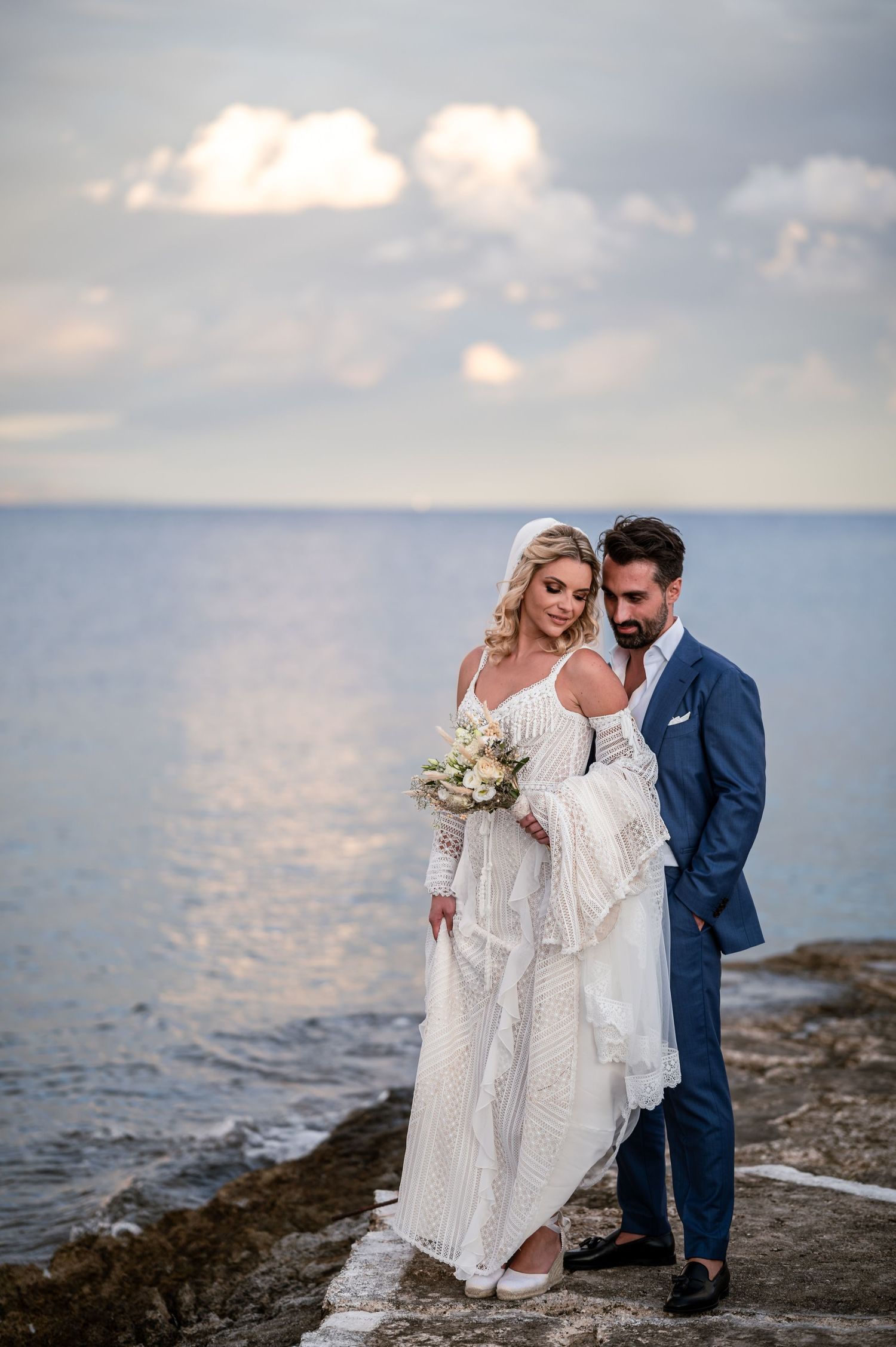 Newlyweds pose together on rocky coastline with ocean and cloudy sky backdrop.