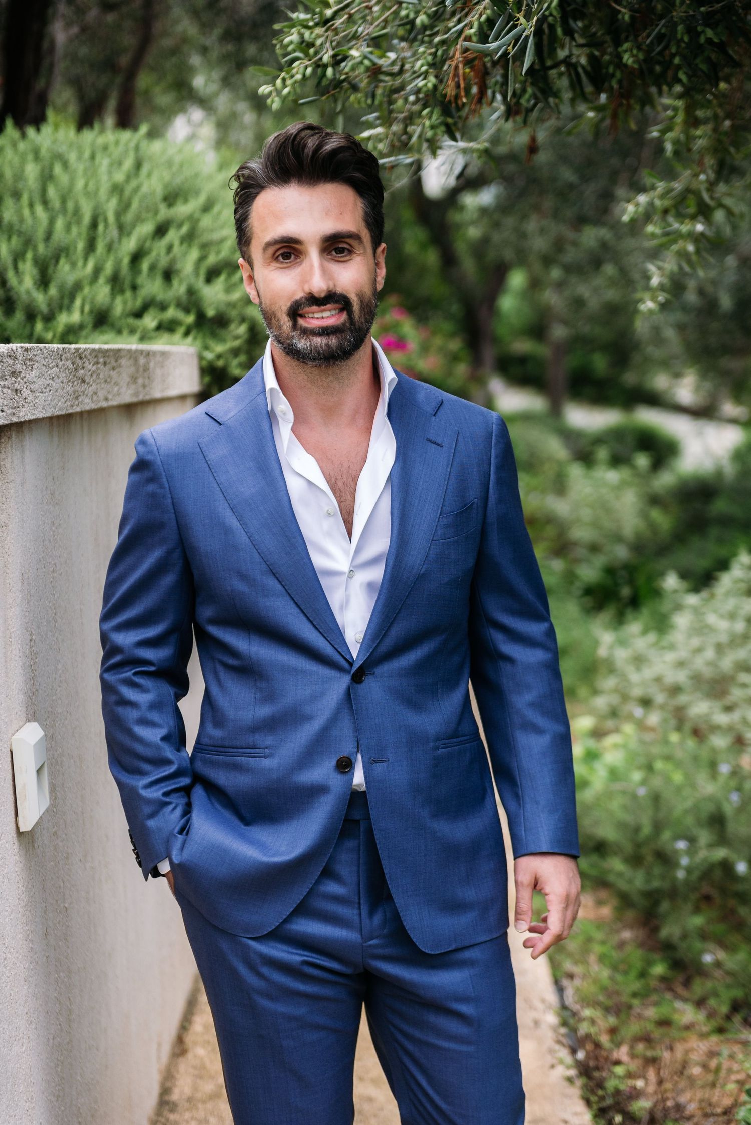 Man walks confidently in tailored blue suit through garden setting.