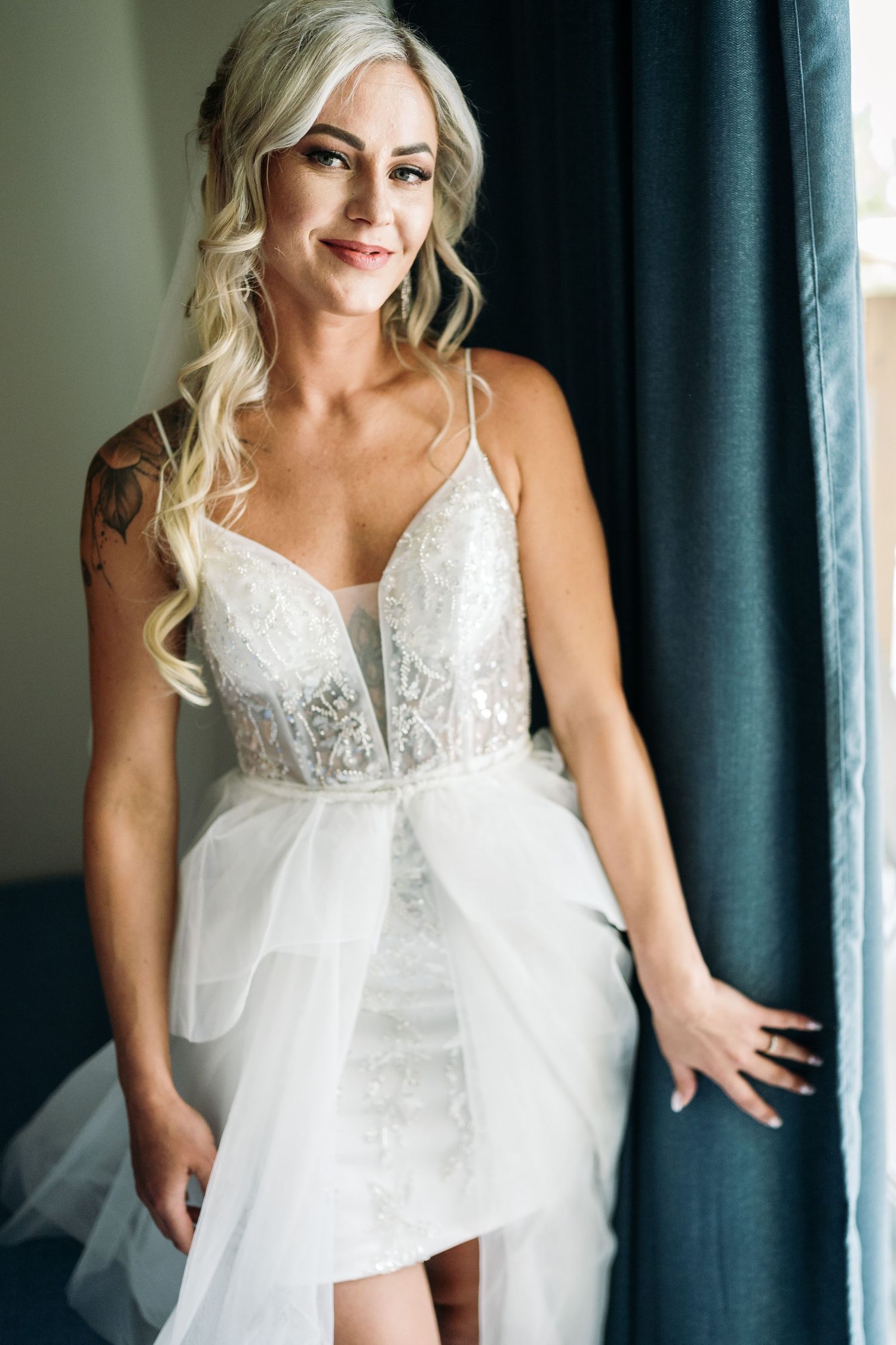 Bride in white lace wedding dress with flowing train poses by window.