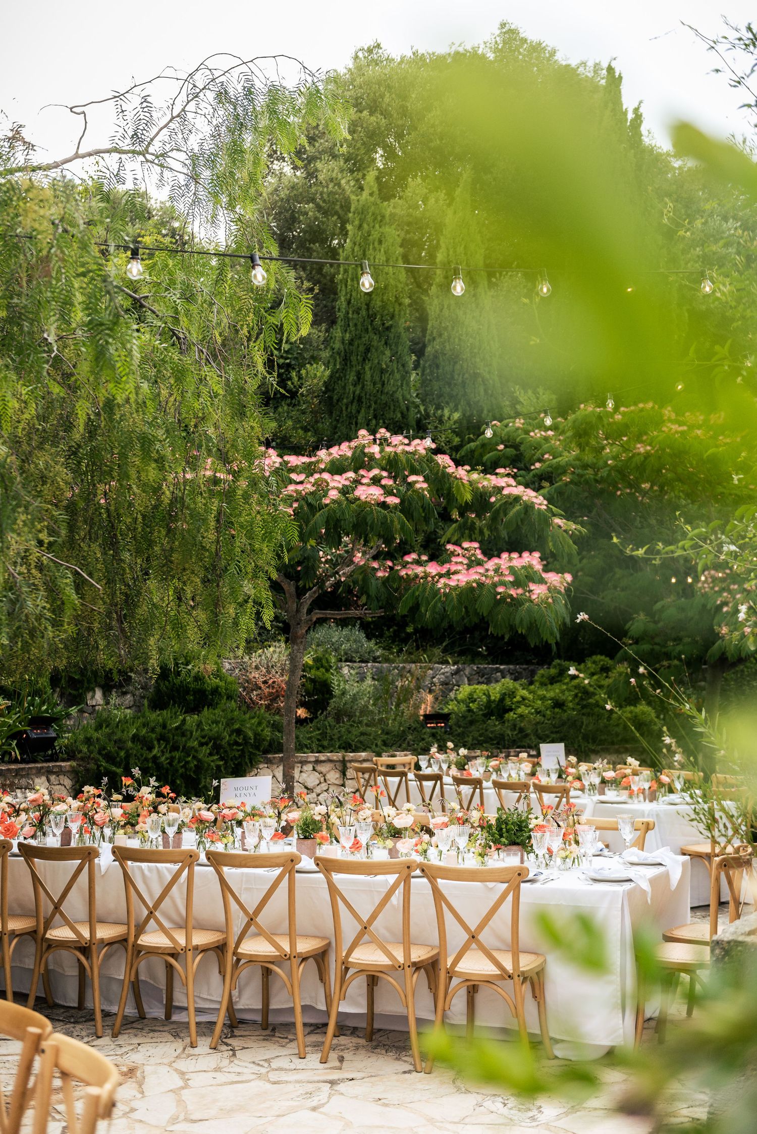 Garden wedding reception setup with wooden chairs and white tablecloths under flowering trees.