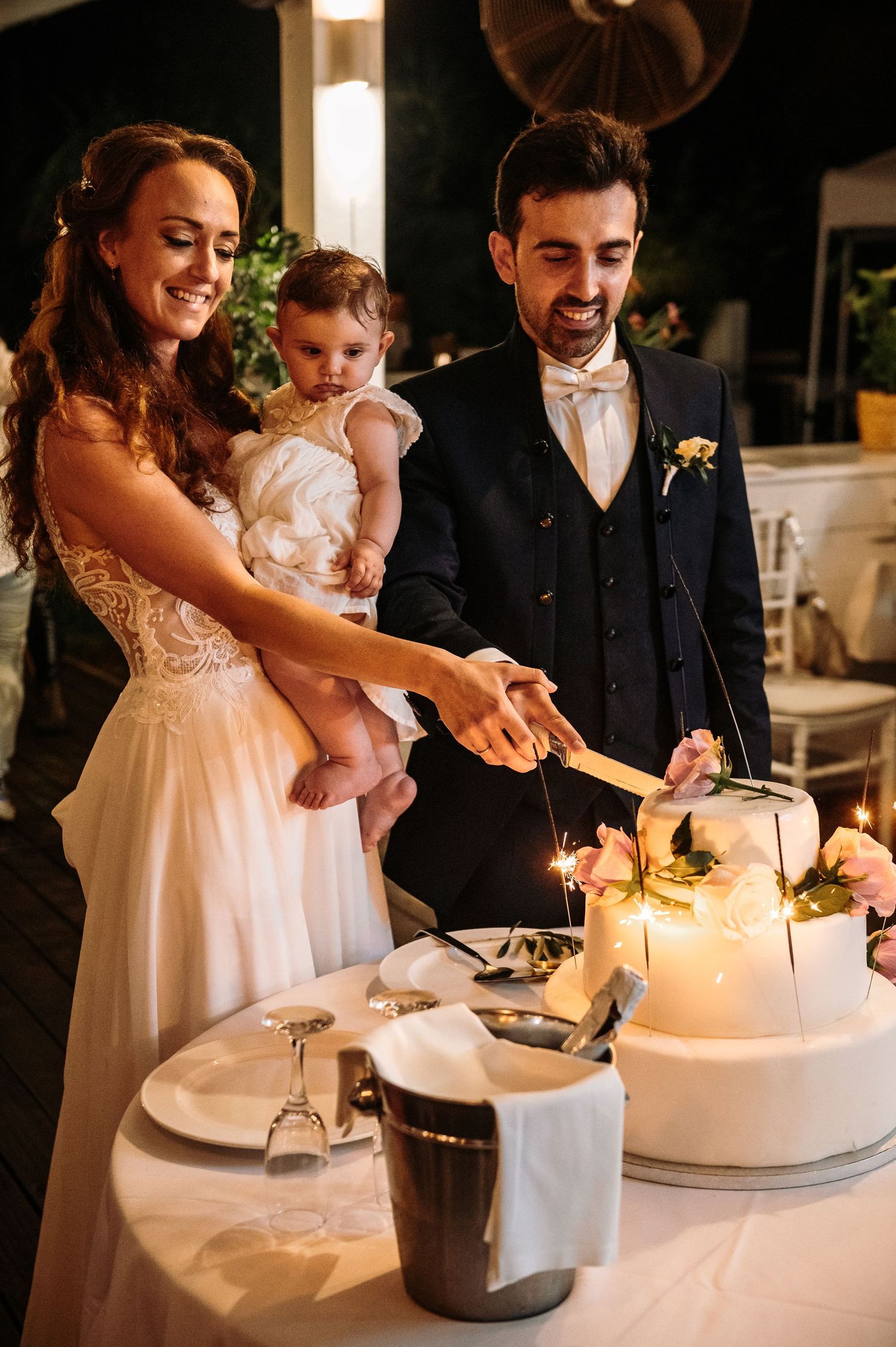 Couple cutting their white wedding cake together during an evening reception.