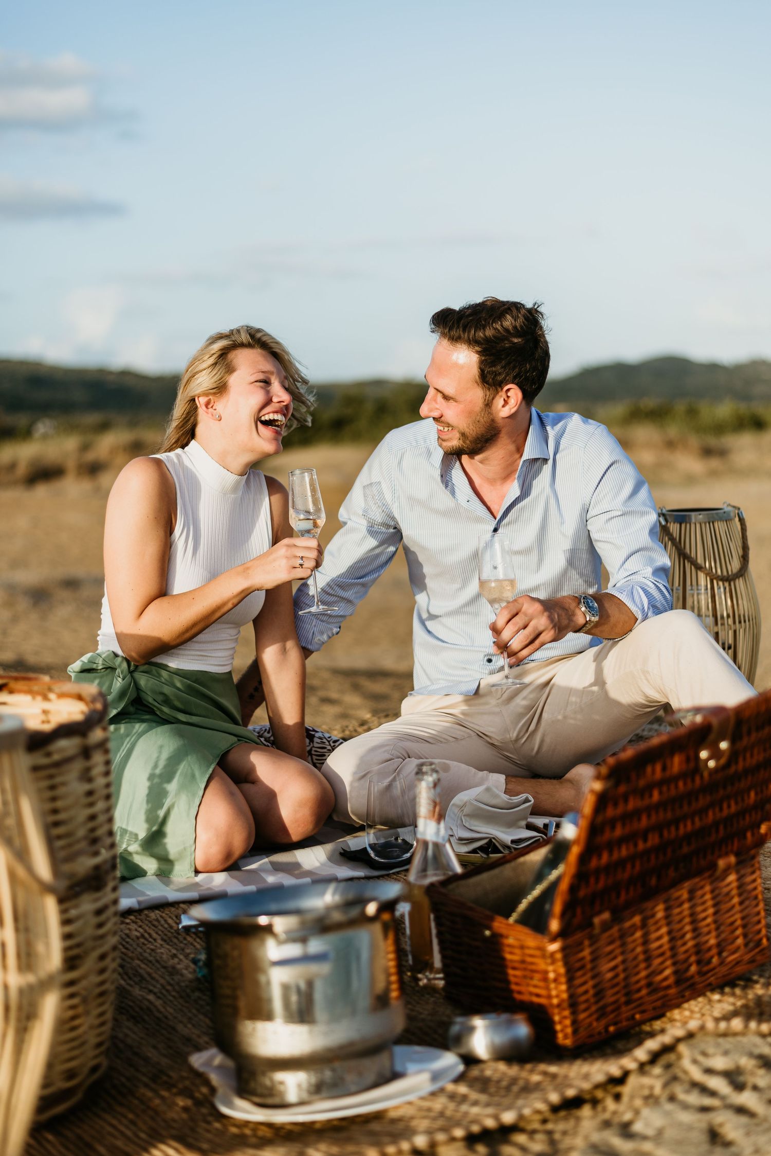 Couple enjoying champagne and laughter during a sunset beach picnic.
