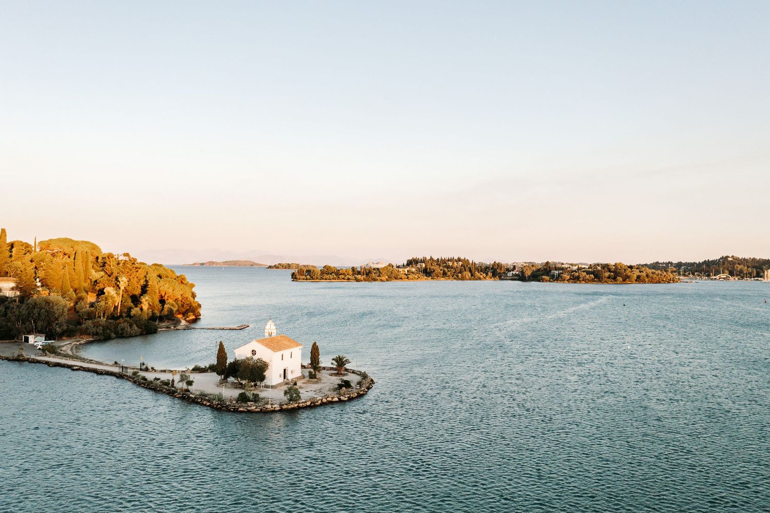 Aerial view of a white church on a small peninsula surrounded by Mediterranean waters at sunset.