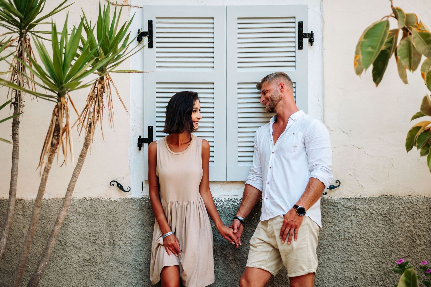 Couple sharing intimate moment against blue shuttered Mediterranean building with tropical plants.