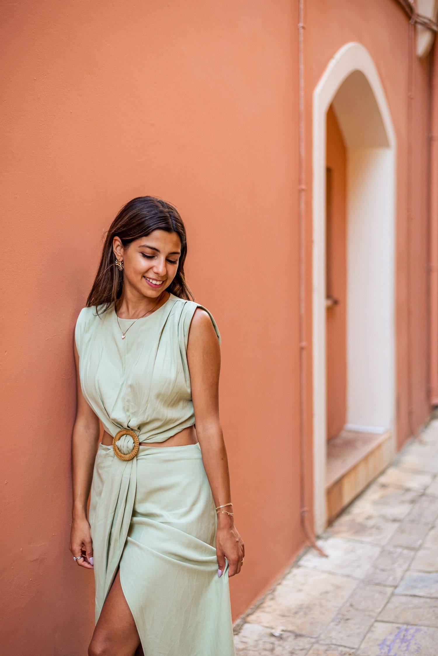 A sage green dress stands out against a coral wall in a Mediterranean street setting.