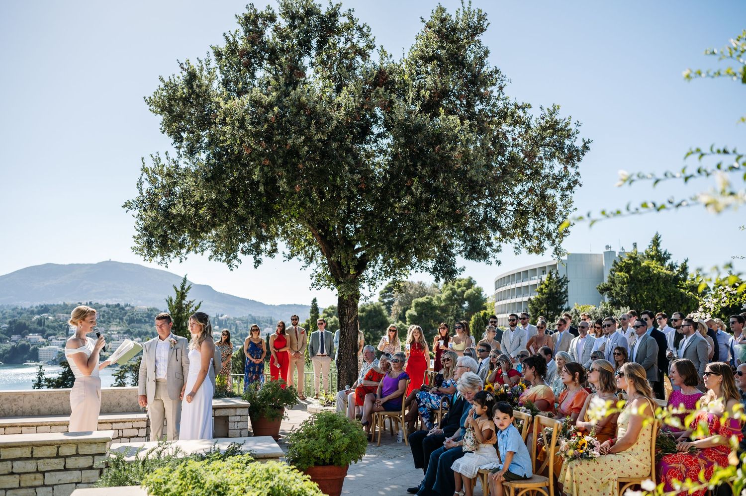Outdoor wedding ceremony with guests gathered under trees with ocean view.