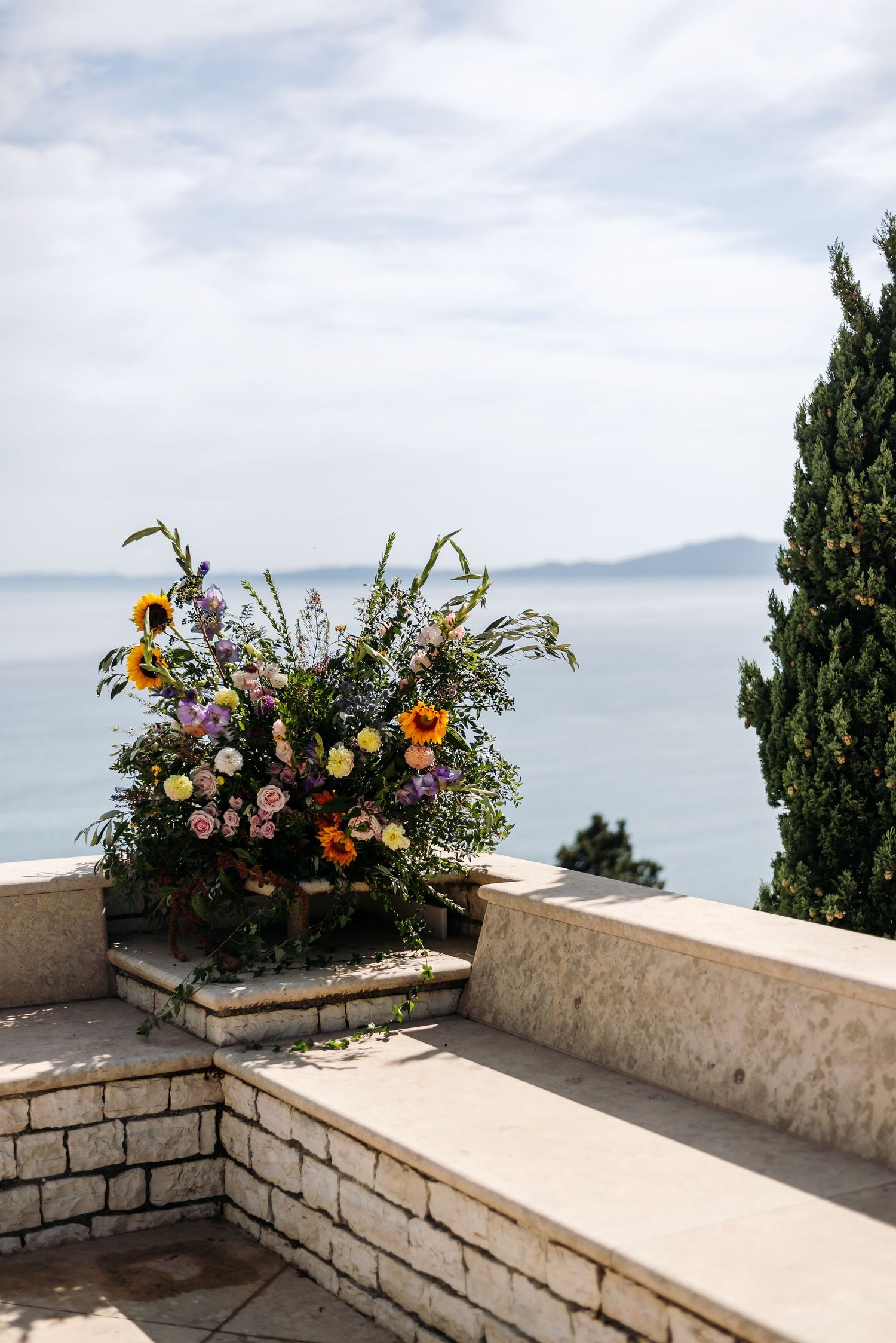 Colorful floral arrangements adorn a stone wall overlooking a serene ocean vista.