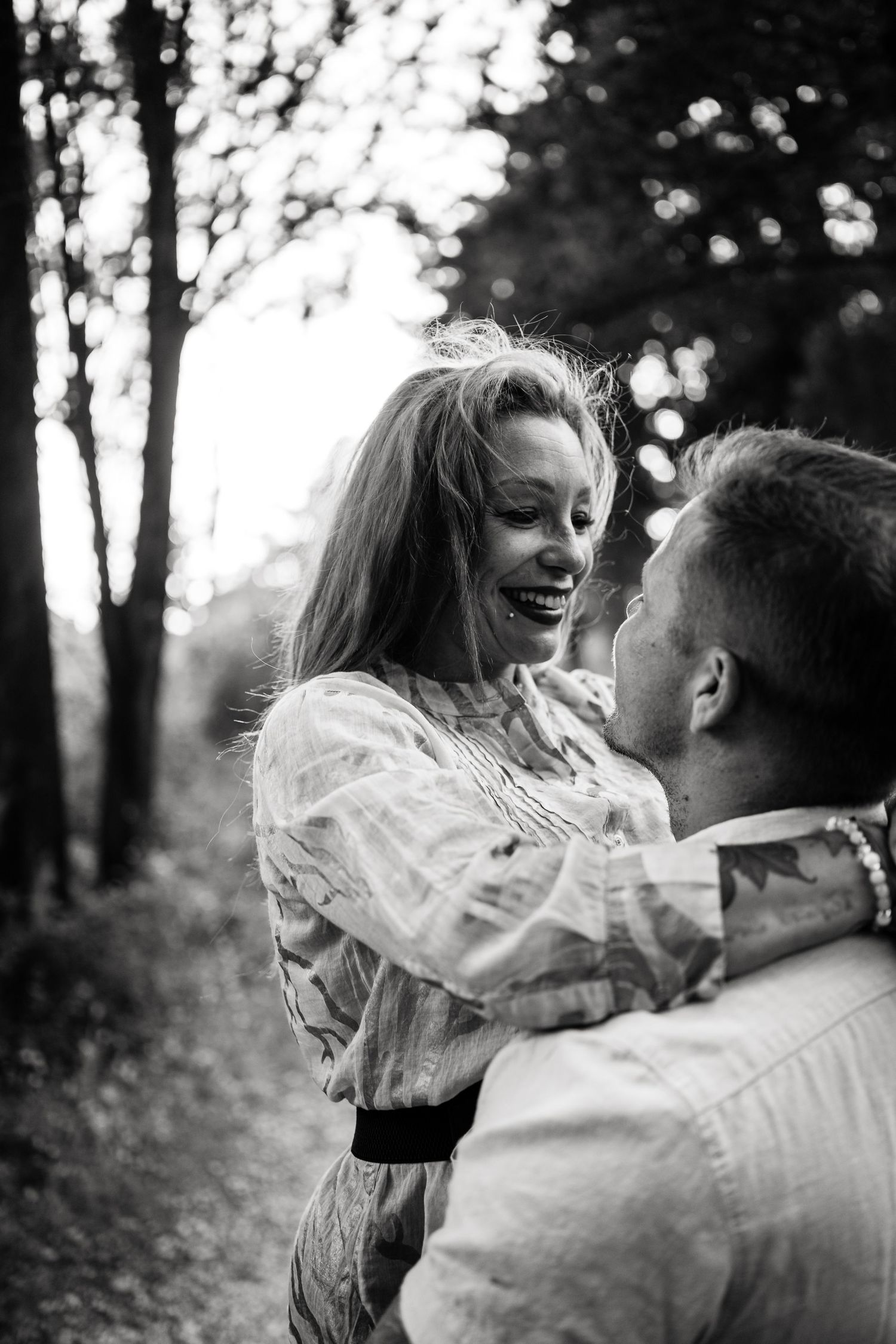 A black and white photo captures a playful moment between a couple in a wooded setting.