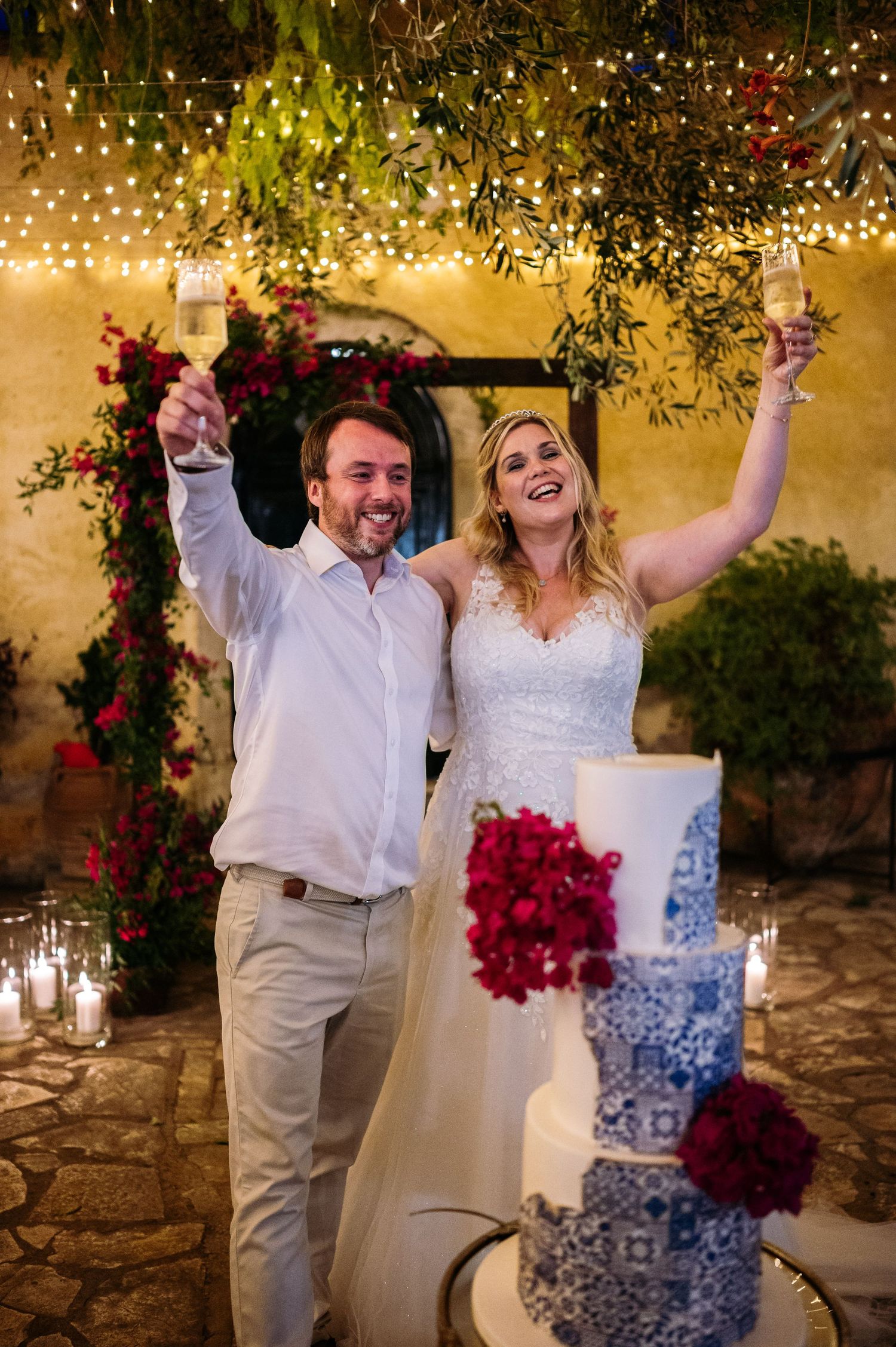 Newlyweds celebrate joyfully with raised glasses beside their ornate blue and white wedding cake.