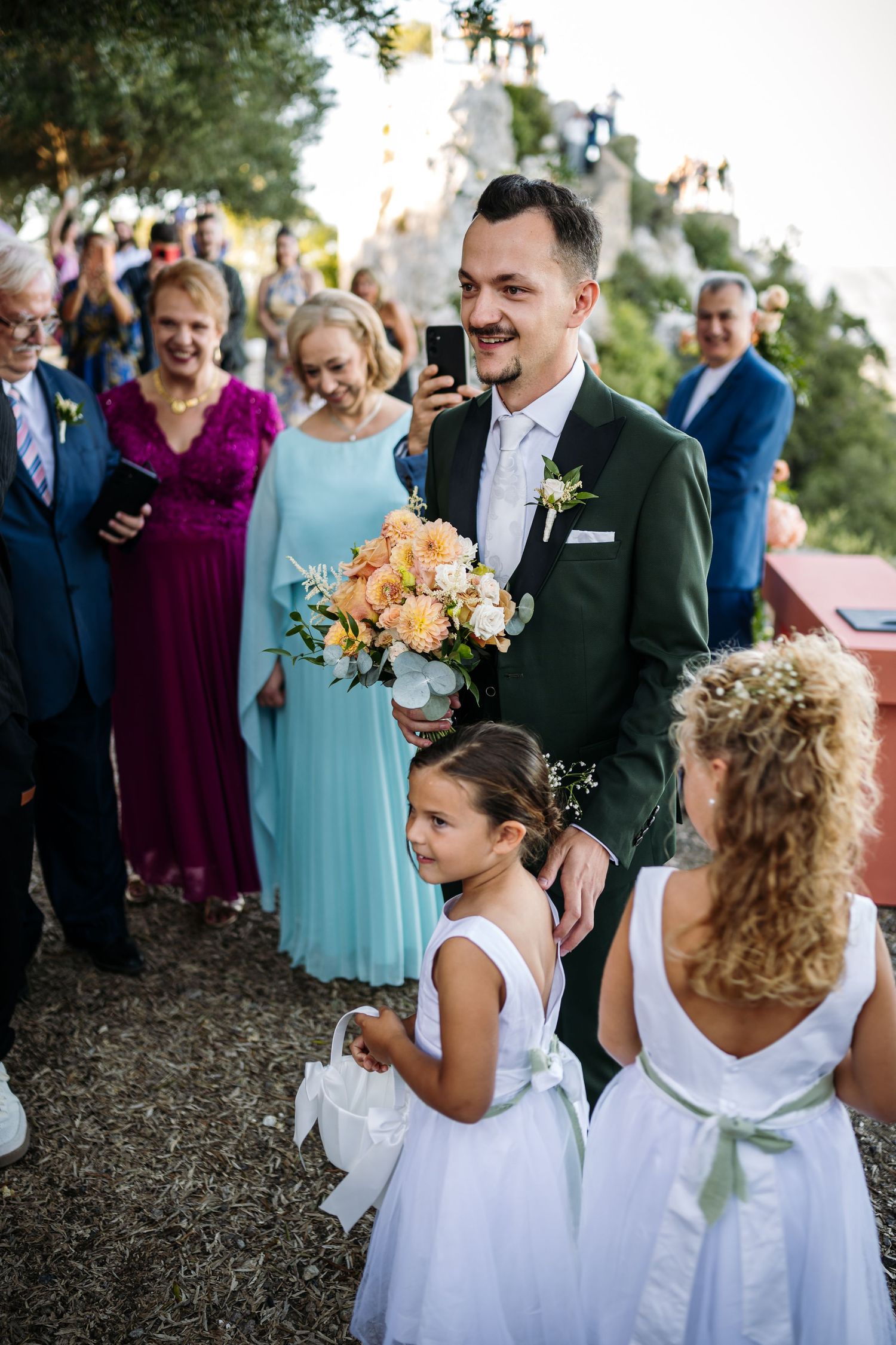 Flower girls in white dresses walk through an outdoor ceremony space decorated with peach floral arrangements.