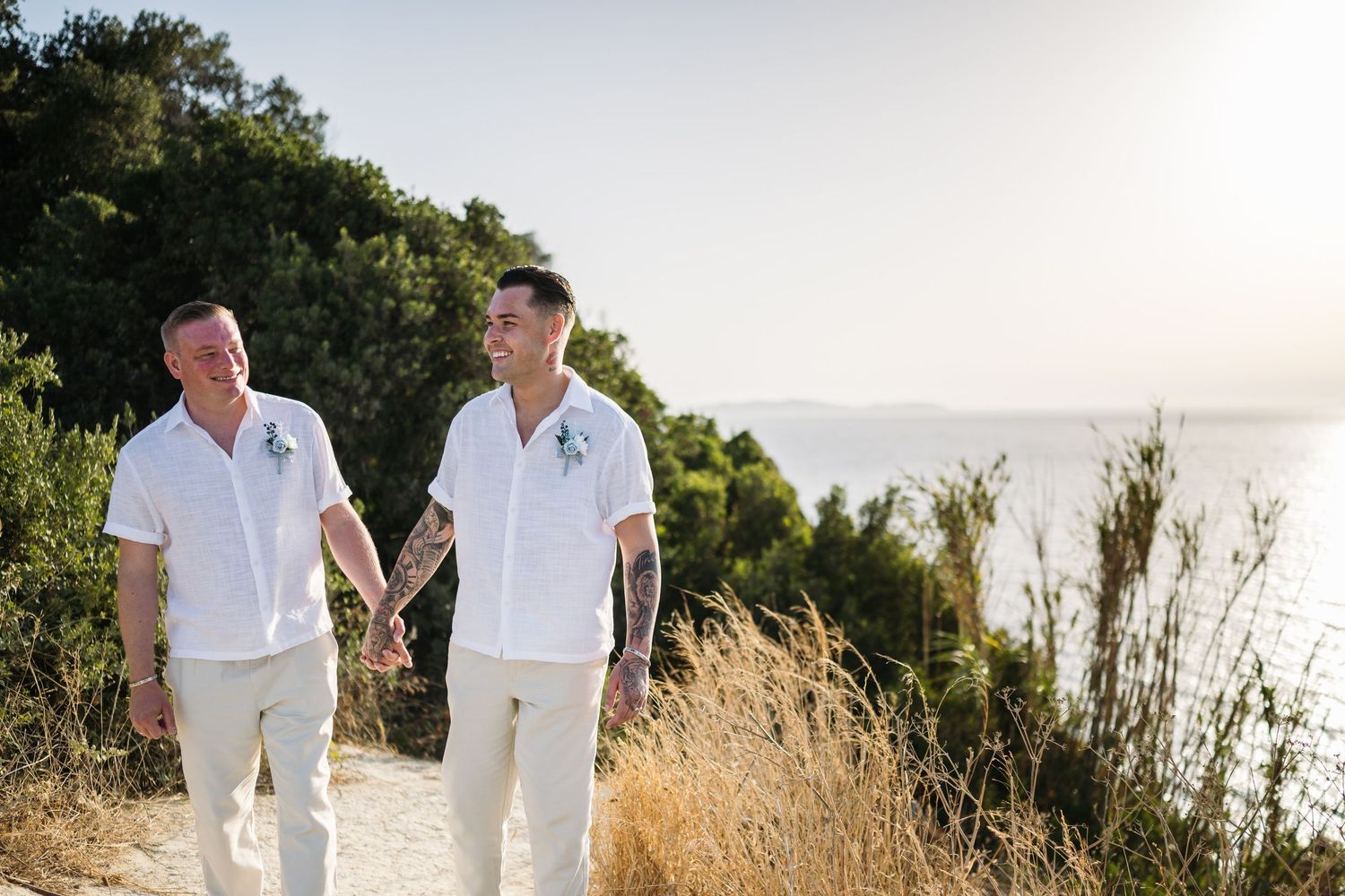 Couple in matching white outfits holding hands while walking along coastal cliff path at sunset.