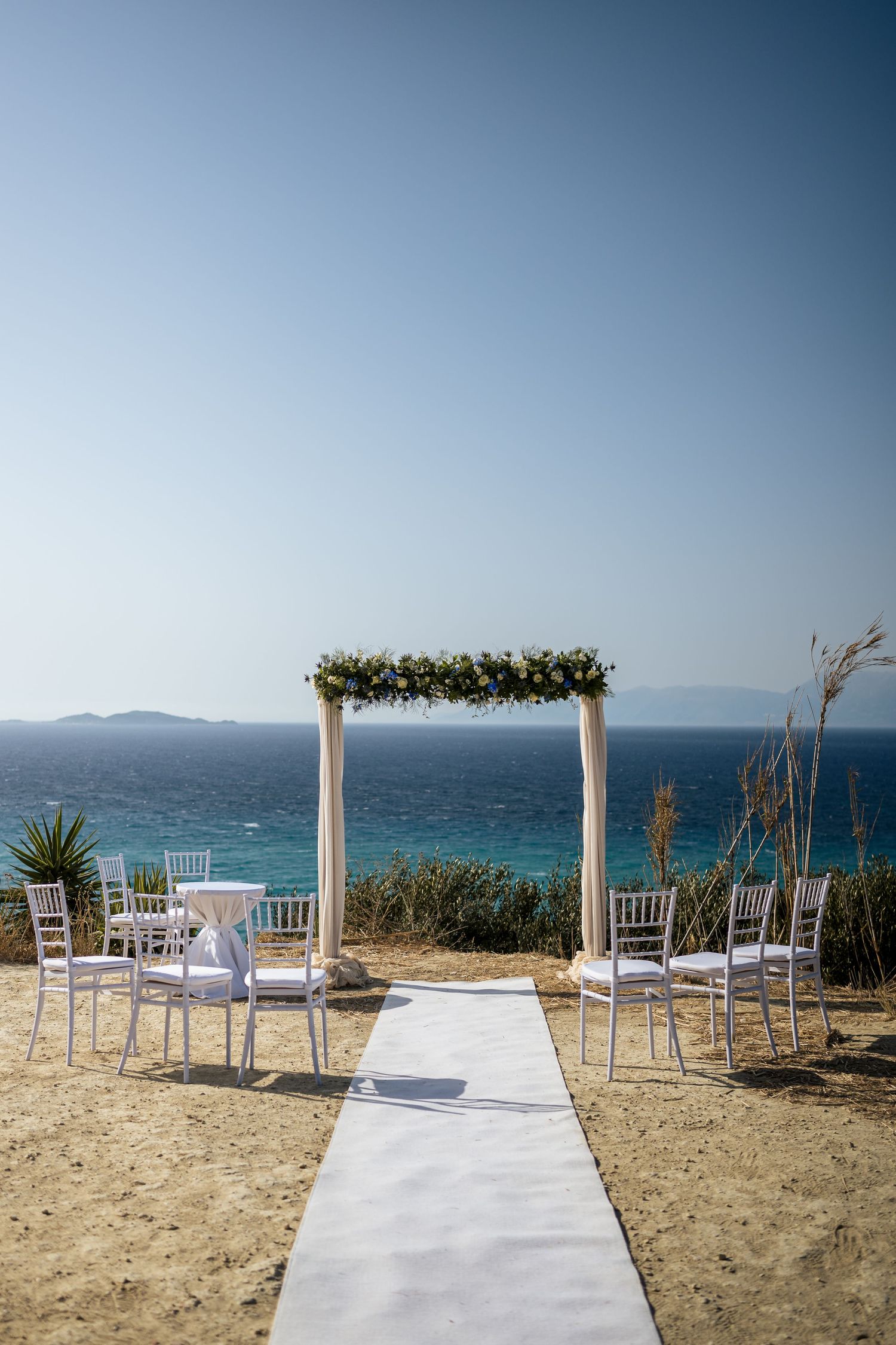 White wedding arch decorated with greenery stands on a beach overlooking turquoise waters and mountains in the distance.