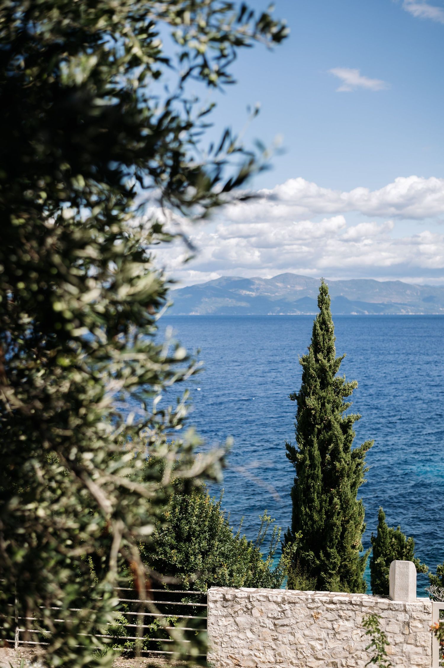 Mediterranean coastal view with cypress trees and stone wall overlooking blue waters and distant mountains.