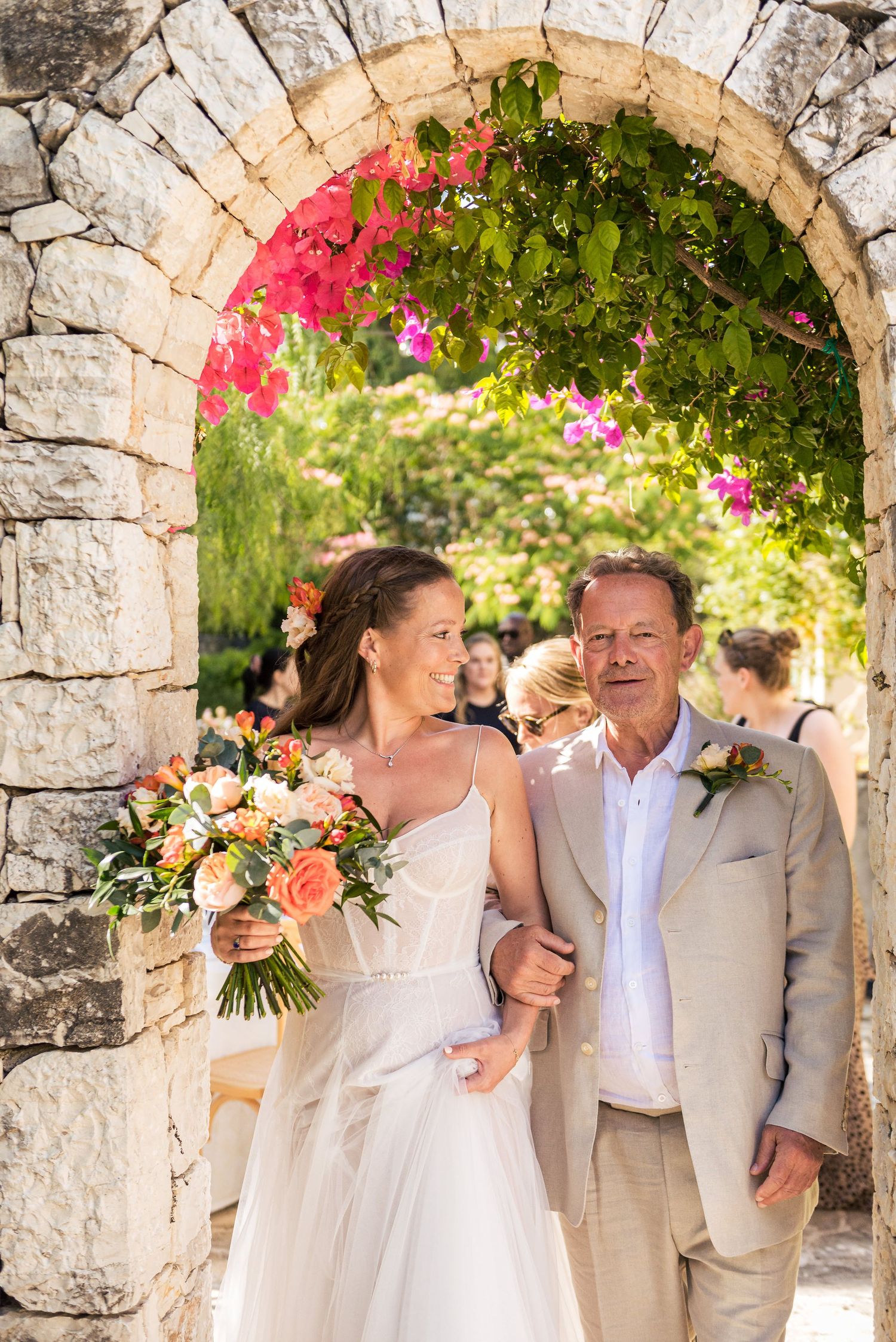 Bride and her father walks through a stone archway decorated with pink bougainvillea flowers in a garden setting.