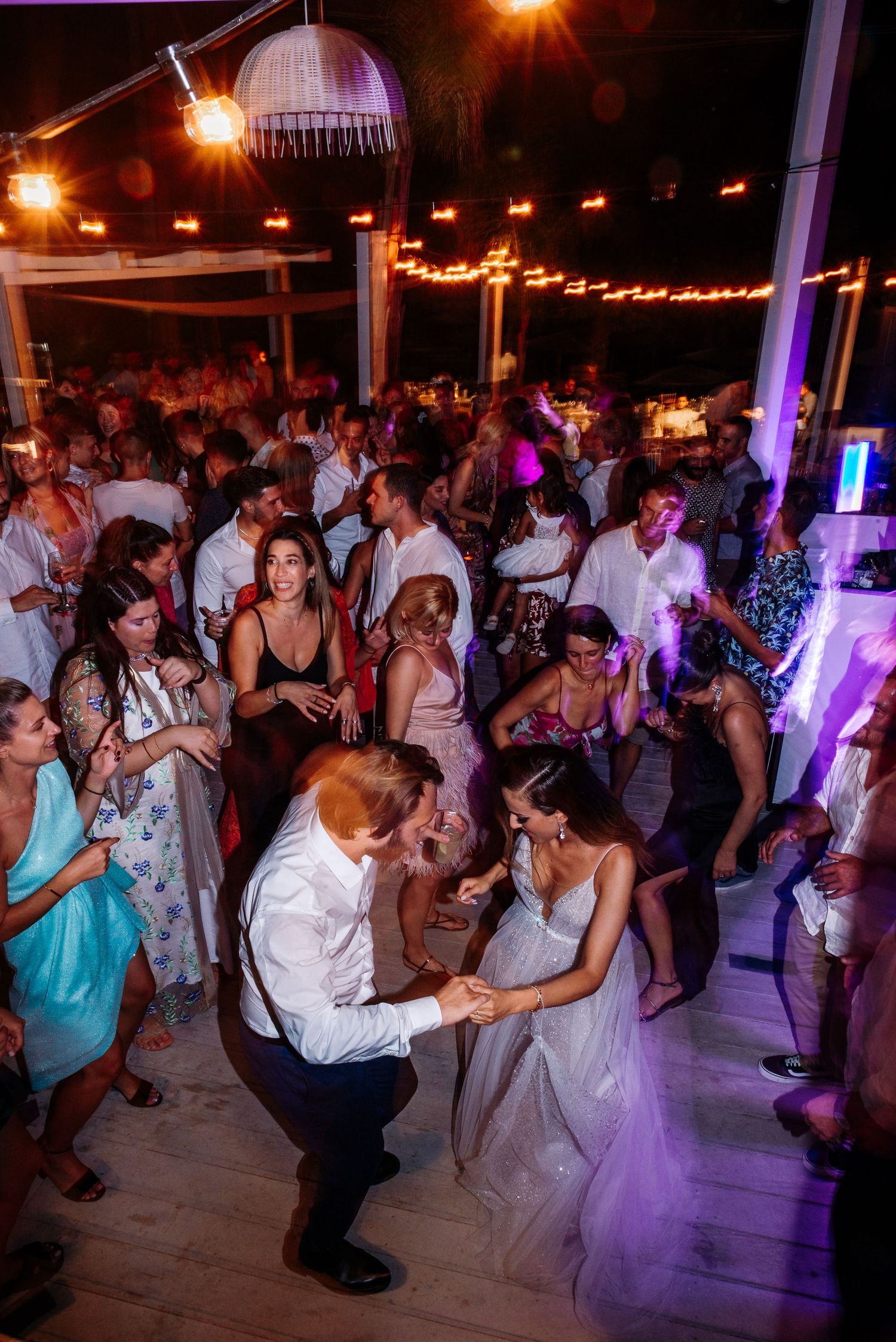 Lively dance floor scene at night wedding reception with purple uplighting and string lights.