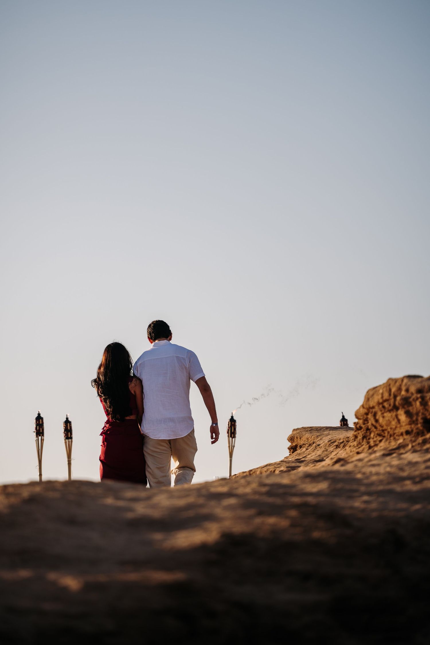 Silhouettes against sunset sky with tiki torches on rocky cliff edge.