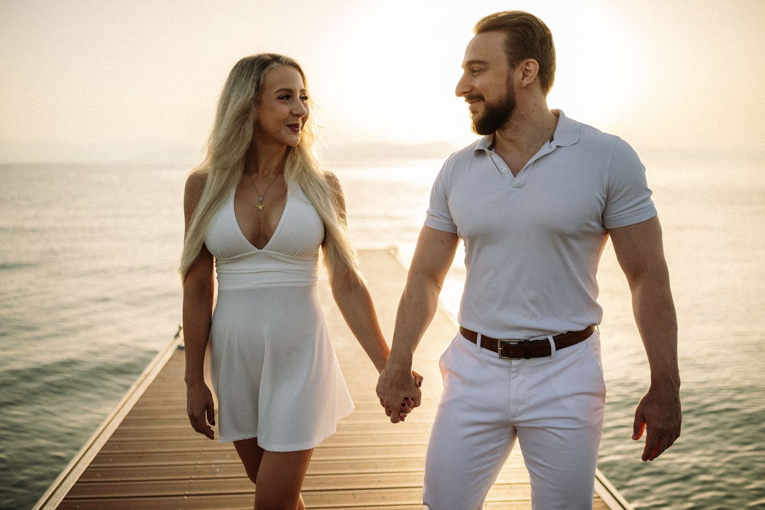 A couple in white outfits walks along a wooden pier at sunset by the ocean.