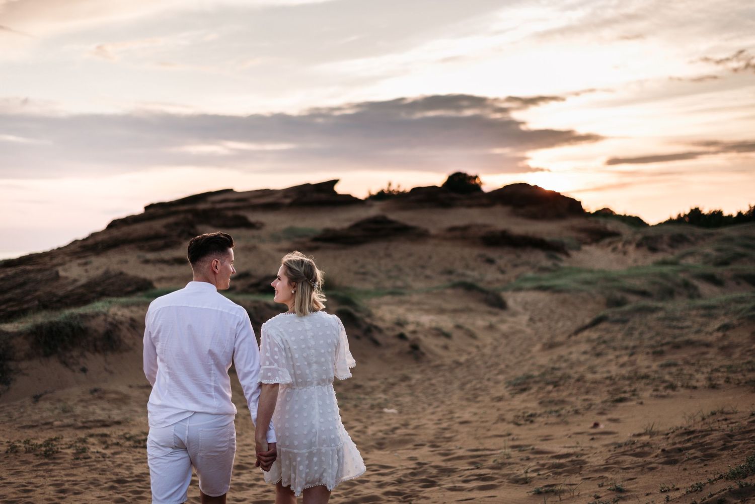 Couple in white outfits walking together on a scenic hilltop during sunset.