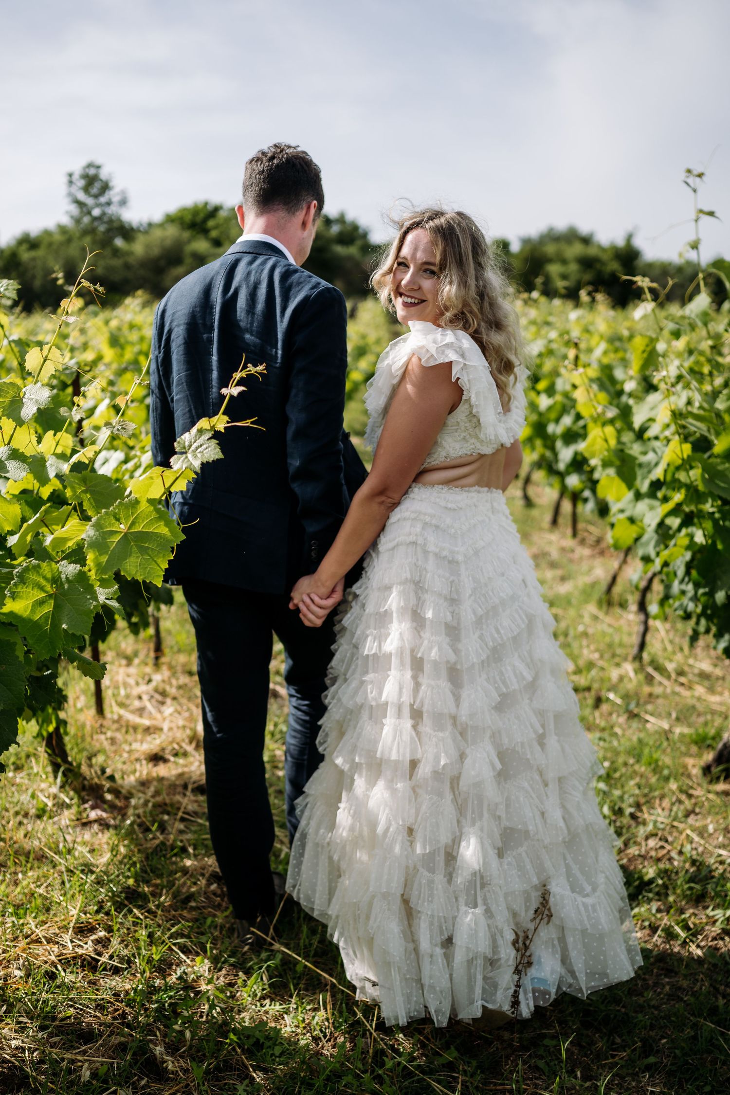 Couple walks through vineyard rows showcasing layered wedding dress.