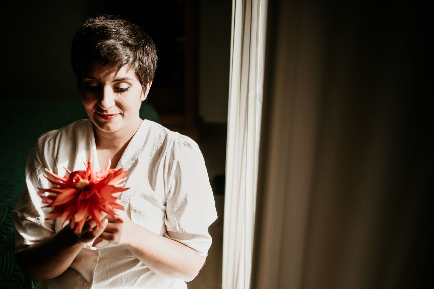 Person in white holding a vibrant red flower near a window.