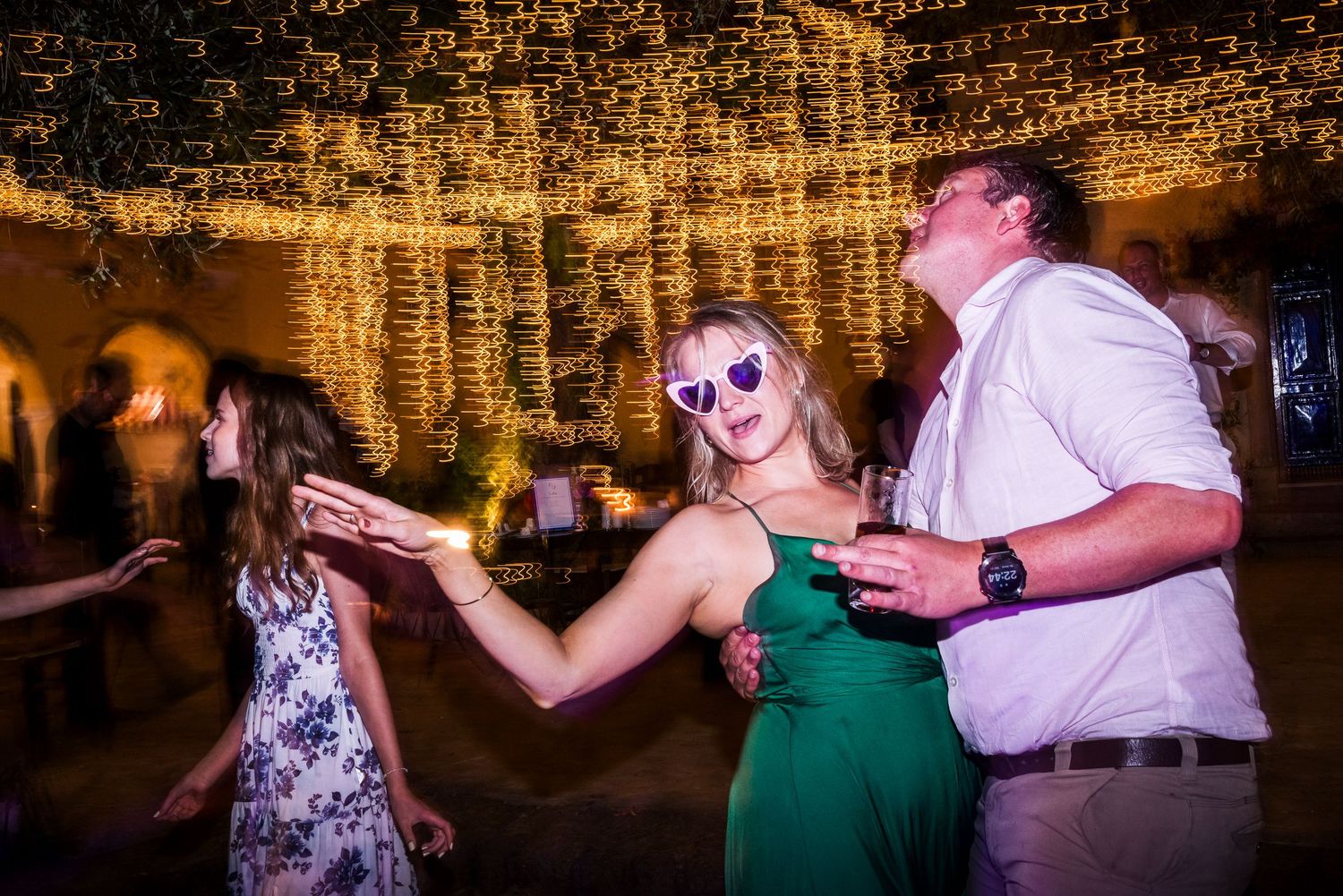 Wedding guests dancing against a warm brick wall backdrop illuminated with string lights.