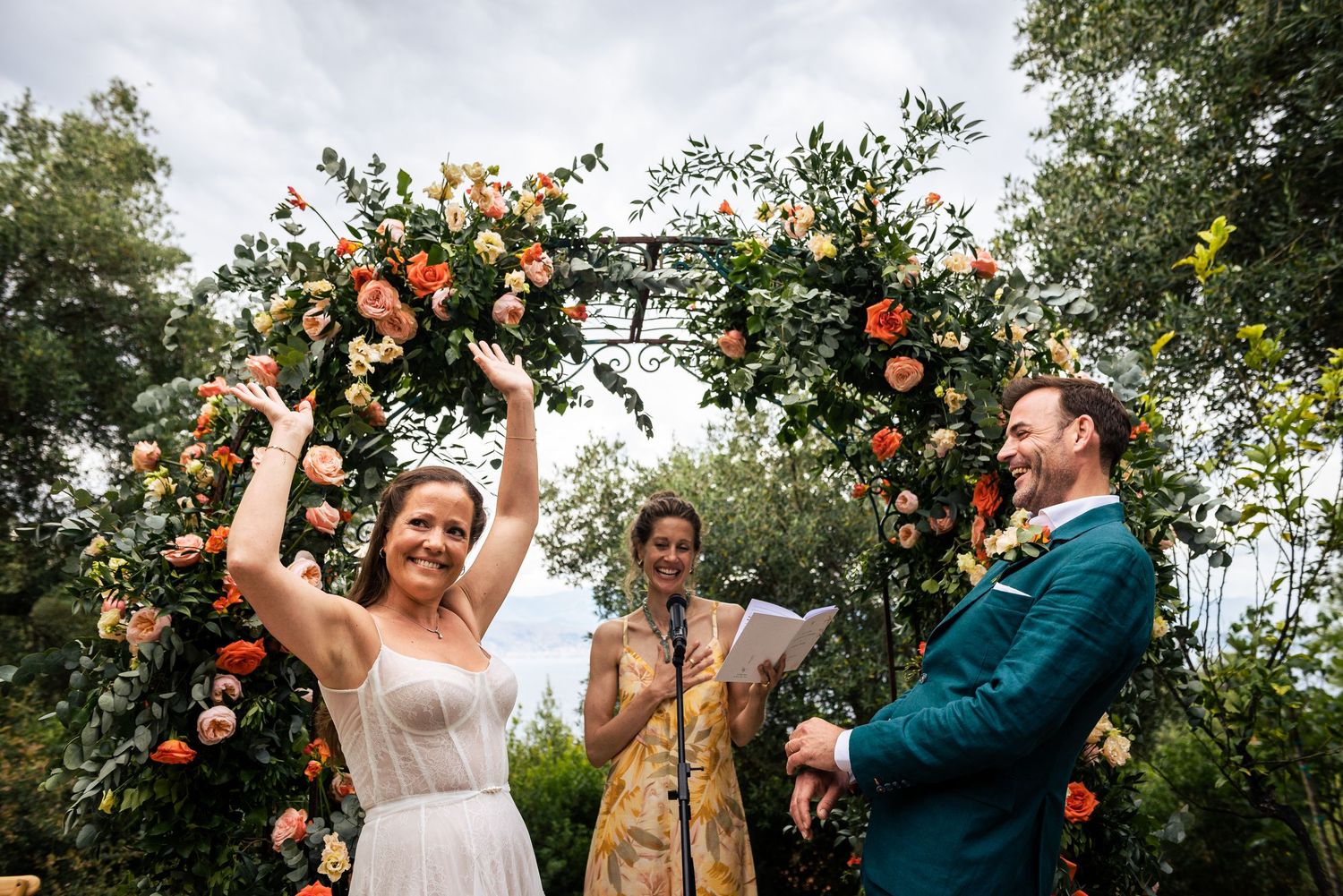 A joyful outdoor wedding ceremony under a floral arch with peach and coral roses.