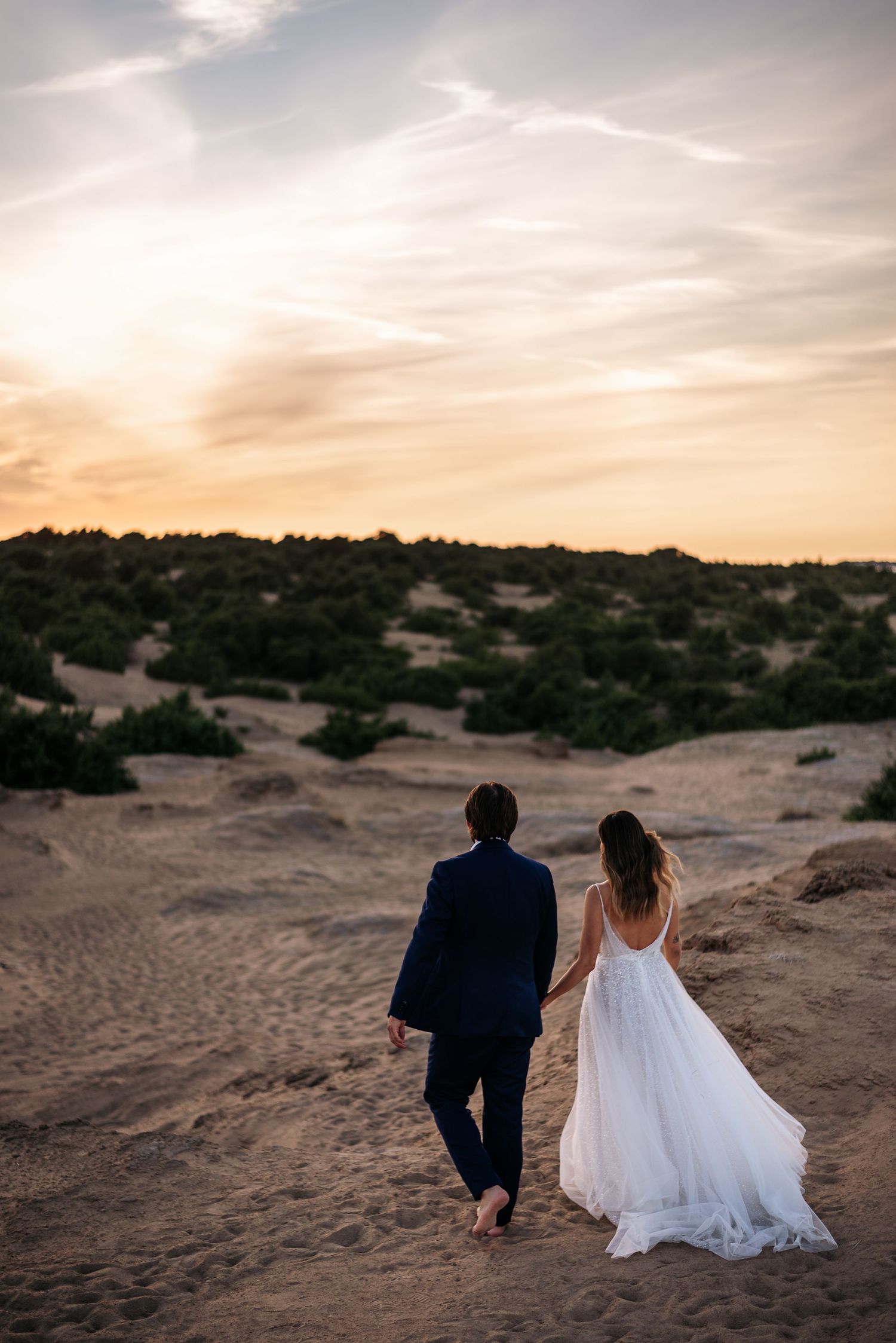Newlyweds walk hand in hand across sandy desert terrain at dusk.
