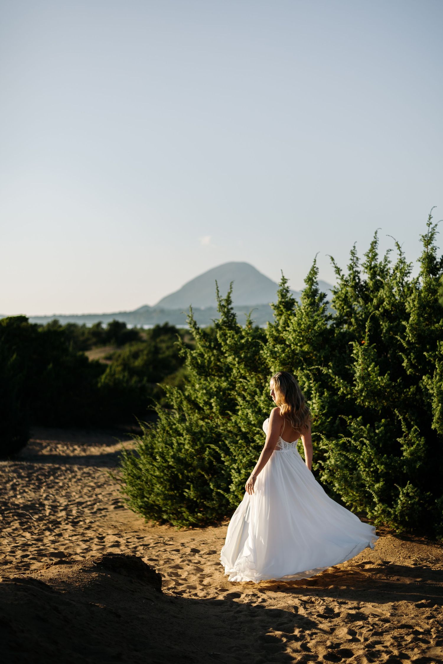 White wedding dress flows gracefully across sandy dunes with mountain peaks in the background.