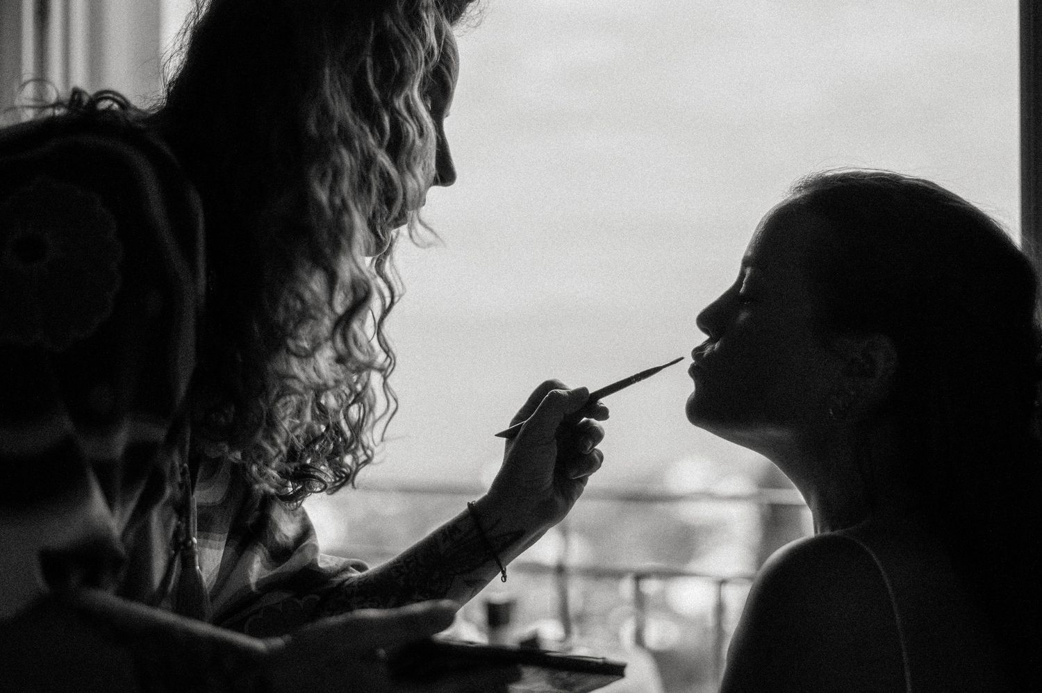 A silhouetted figure applies makeup to another person's face against a bright window backdrop.