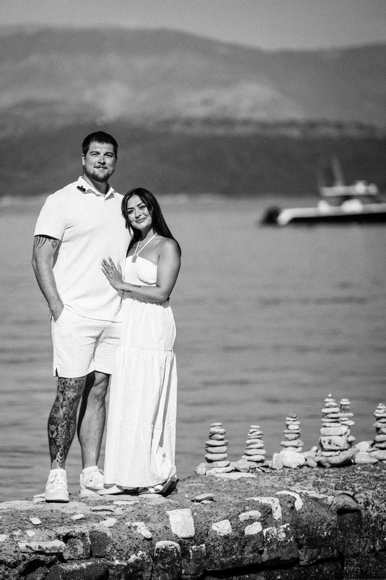 Black and white photo of couple standing on rocky shore with stacked stones.