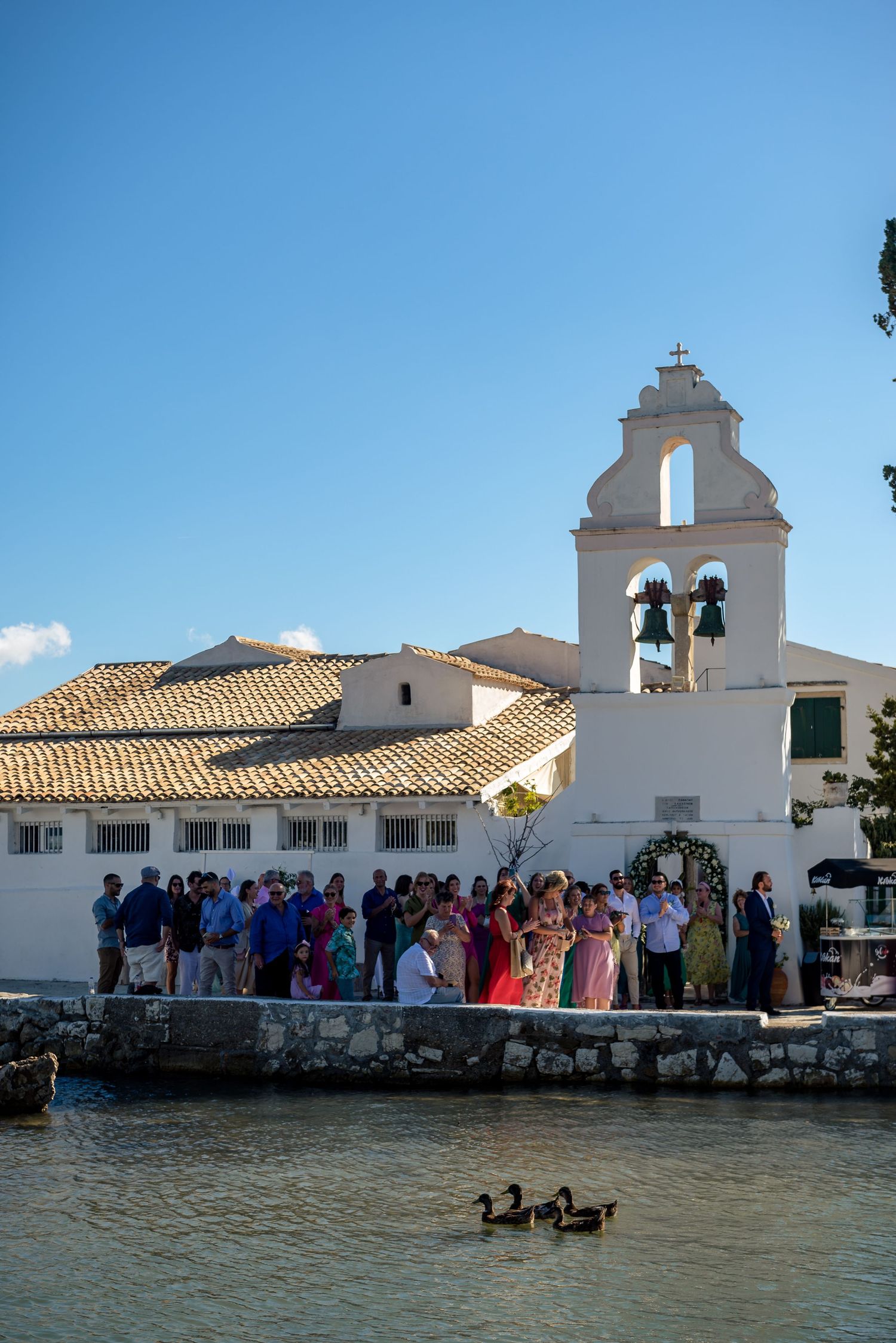 A white church building with bell tower sits along a waterfront with guests gathered outside.