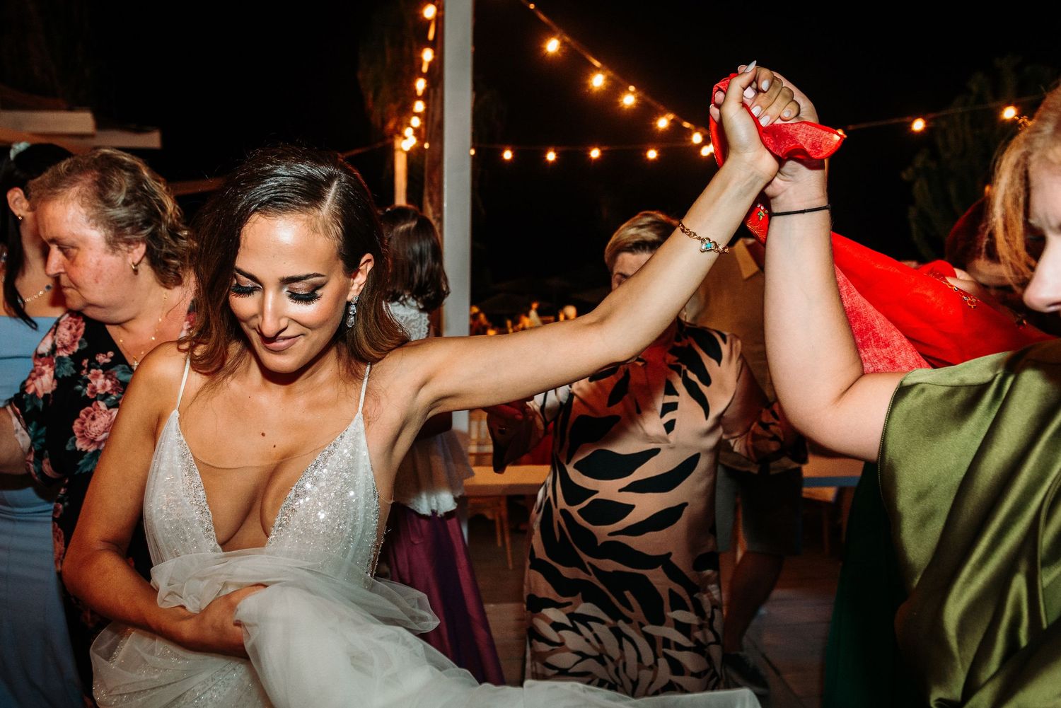 Wedding guests dancing and celebrating with raised arms under string lights at outdoor reception.
