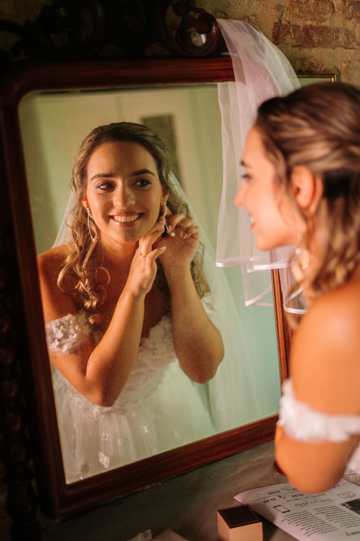 A bride admires her reflection while putting on earrings in an ornate mirror.