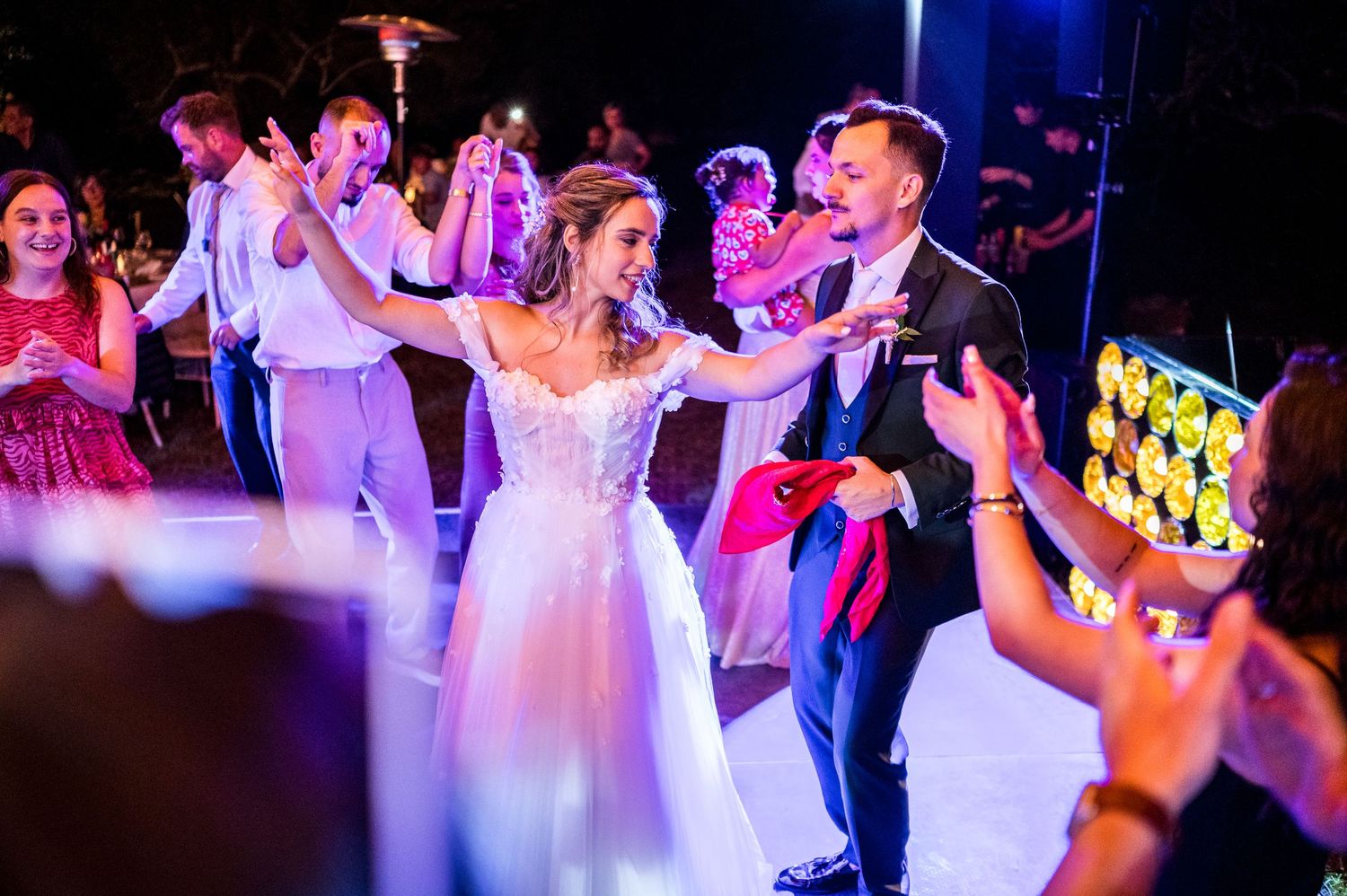 Wedding party dancing under colorful lights during reception celebration.
