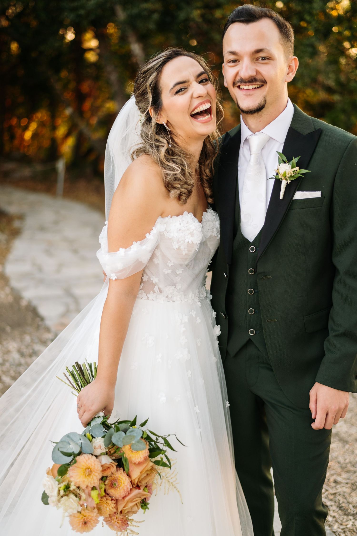 Joyful wedding couple sharing a laugh while holding a peach-colored bouquet outdoors.