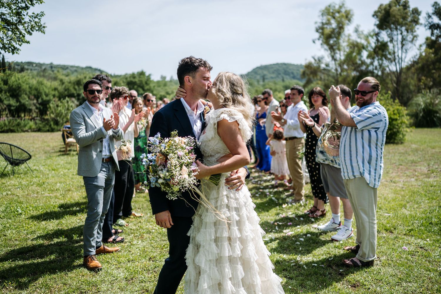 Wedding guests celebrate outdoors with champagne toasts on a sunny hillside.