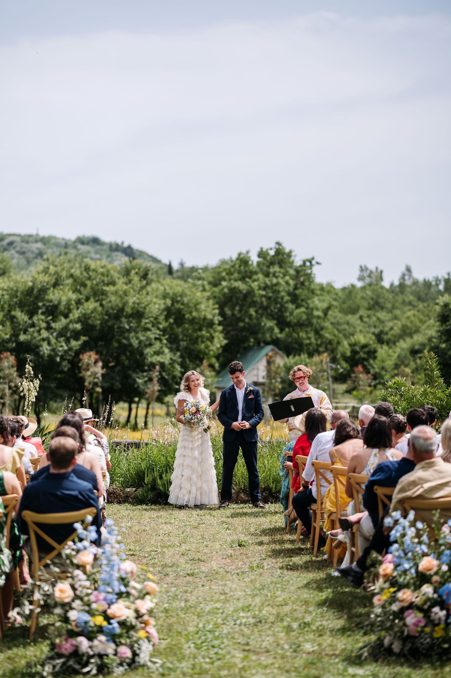 Garden wedding ceremony with floral arrangements and wooden chairs set against rolling hills.