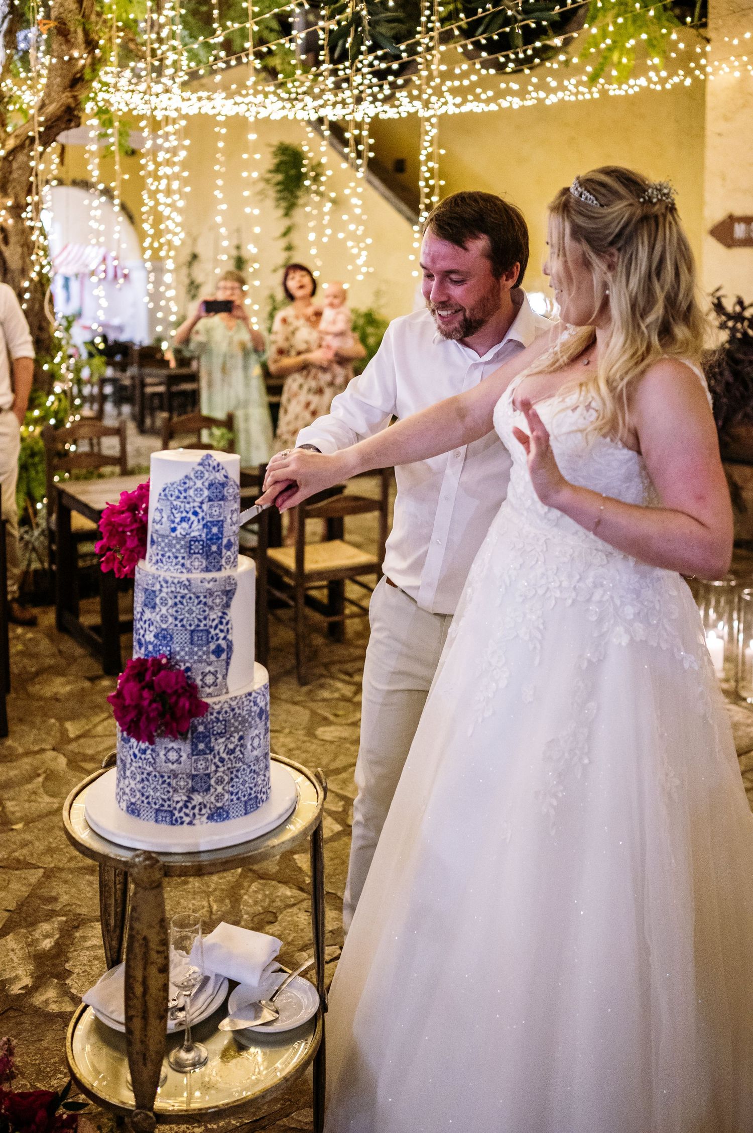 Couple cutting an elegant blue and white wedding cake decorated with red flowers under twinkling string lights.