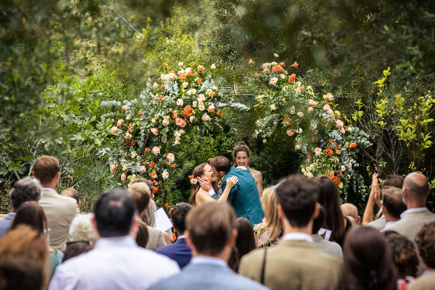 Wedding guests seated outdoors watching ceremony beneath blooming rose bushes.