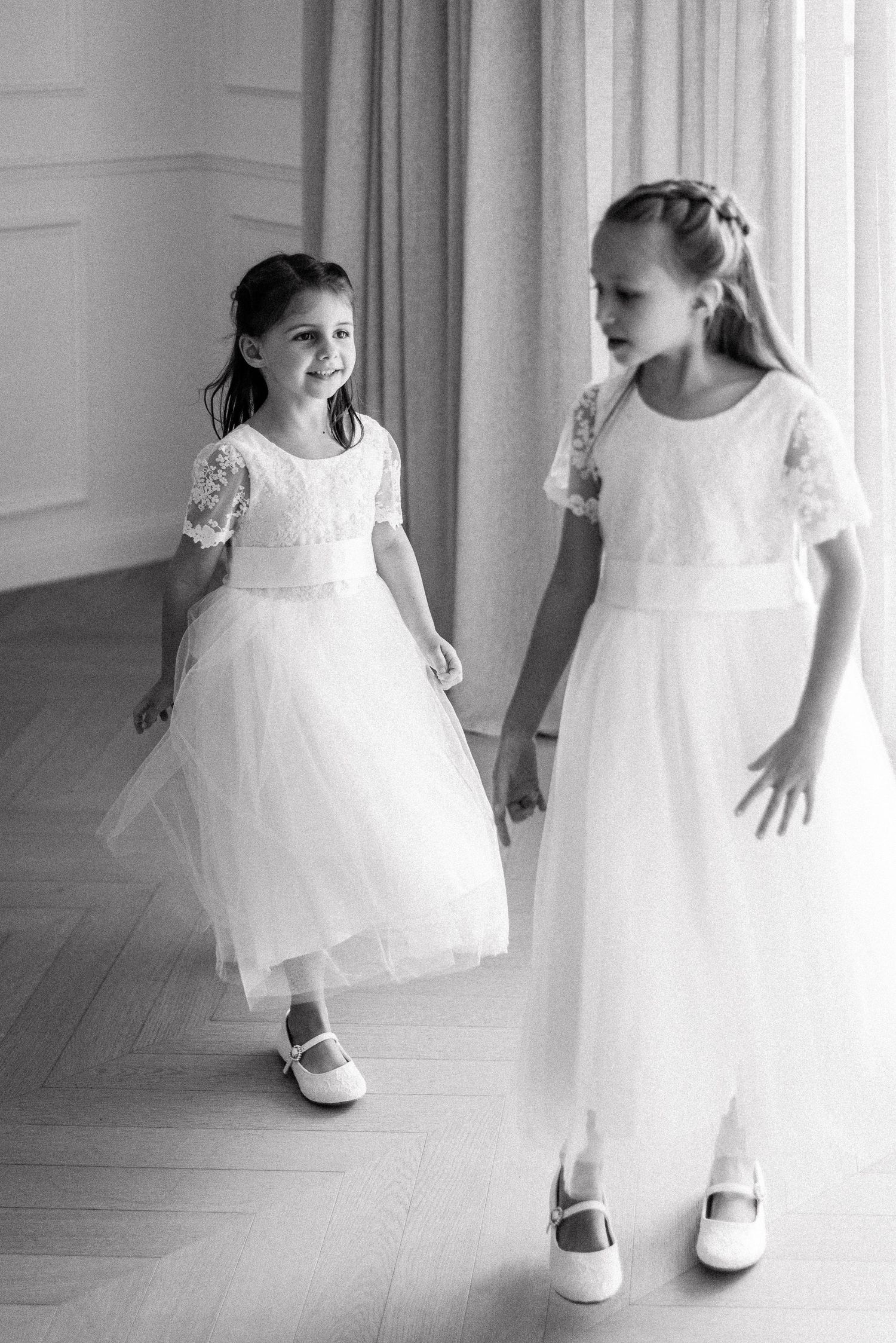 Two young flower girls in white lace dresses and mary jane shoes dancing in an elegant room.