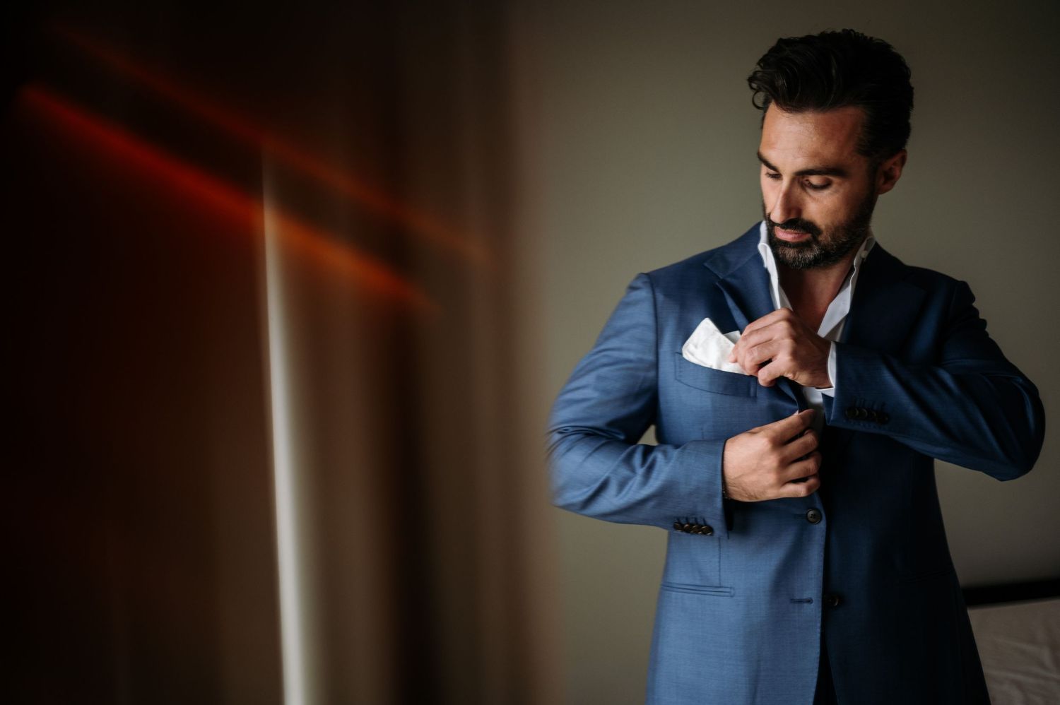 Man in blue suit adjusts his cuffs in moody lighting.