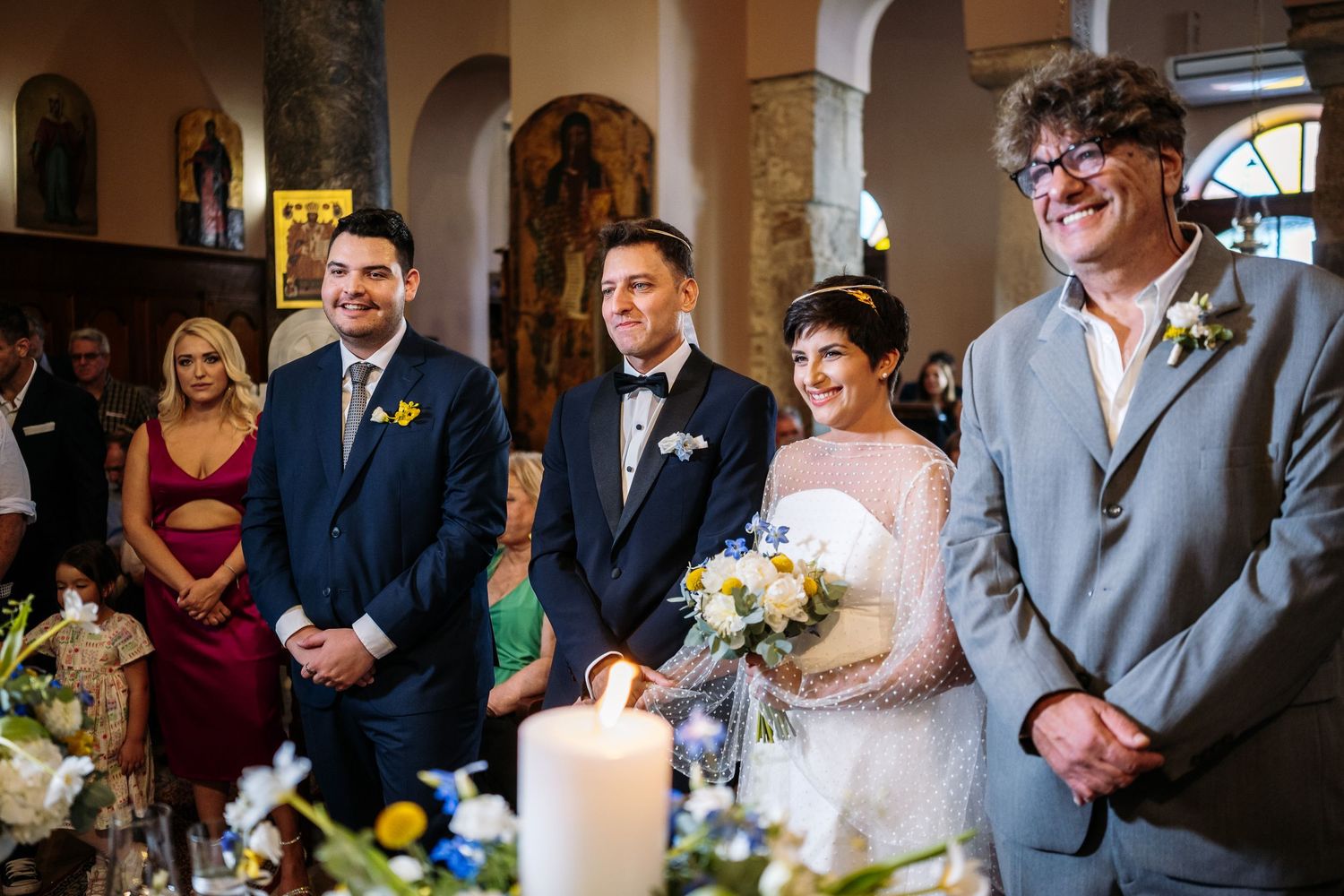 Wedding ceremony attendees gathered in an elegant church interior with arched columns and candlelit decorations.