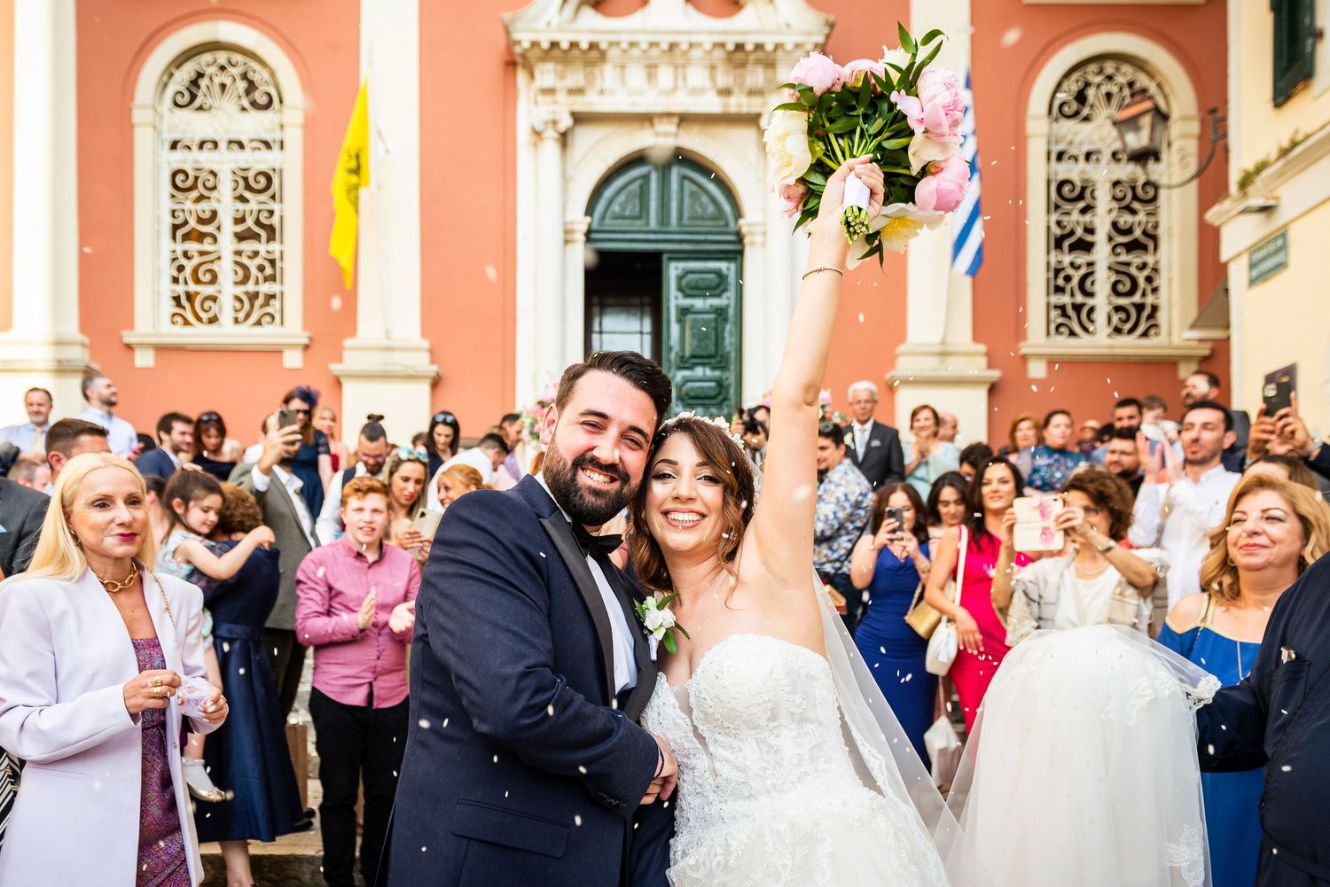 Wedding celebration outside a coral-colored building with ornate architectural features.