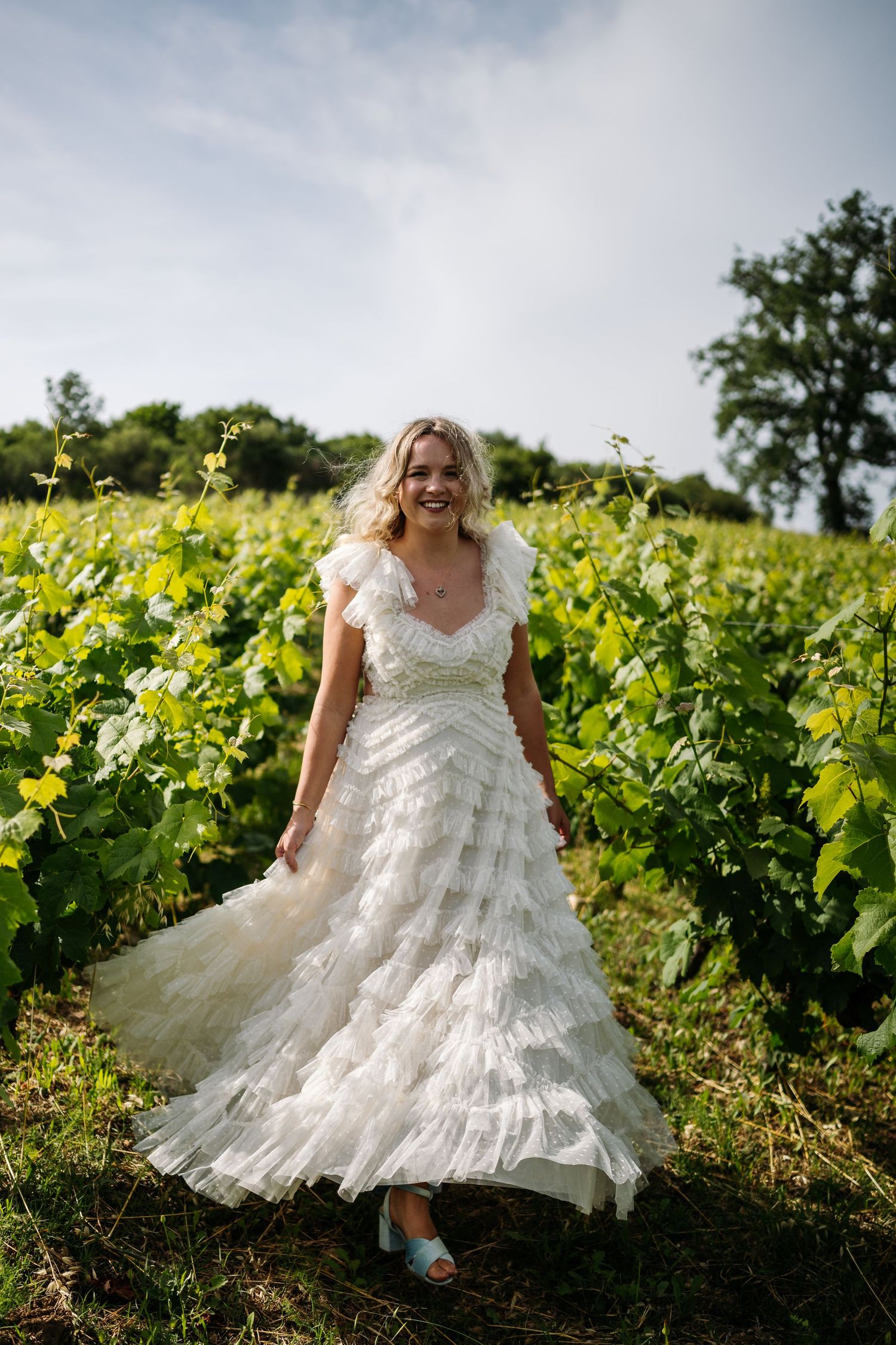 Bride in ruffled wedding dress twirls through sunny vineyard.