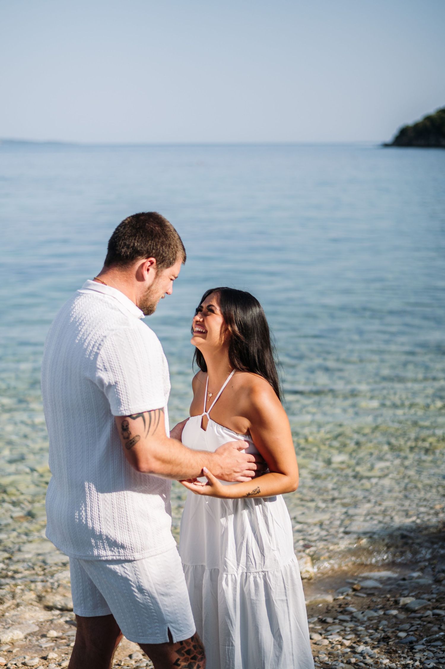 Couple in white outfits sharing a joyful moment by the ocean.