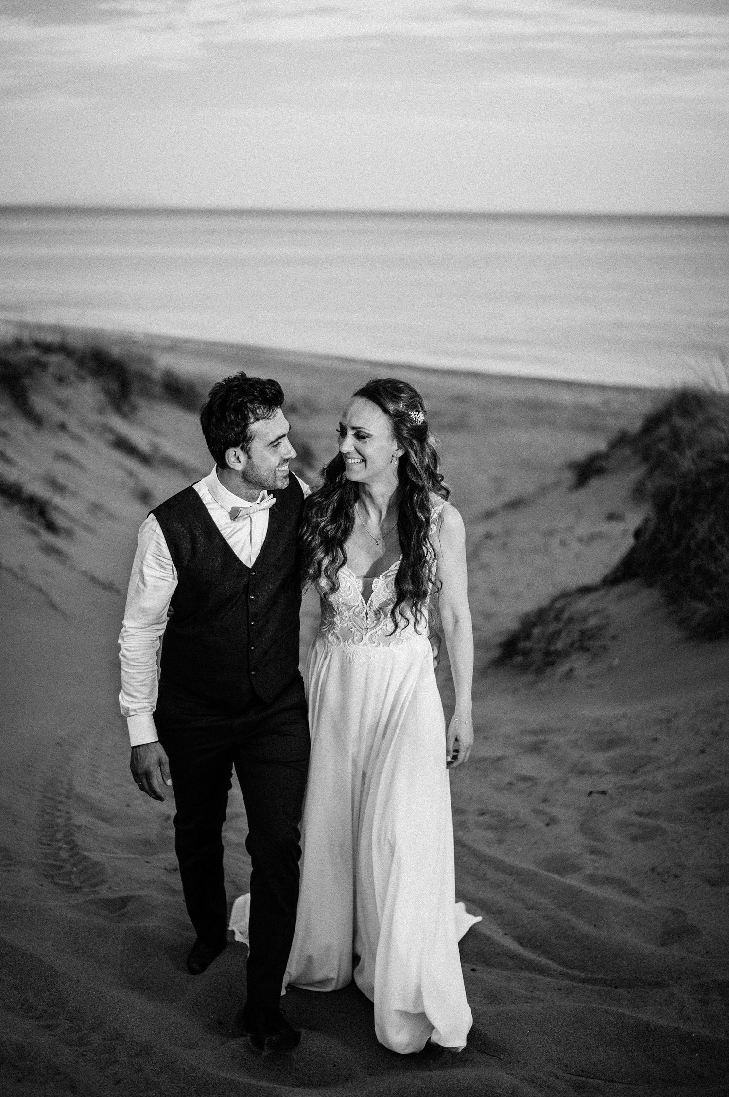 A couple in wedding attire walks together on sand dunes during a black and white beach photoshoot.