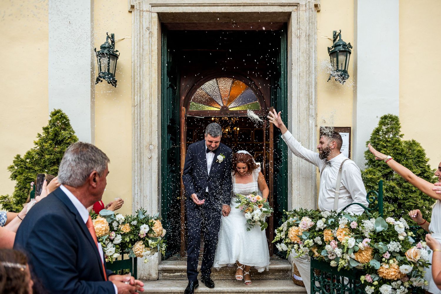Wedding party exits church through ornate doorway decorated with floral arrangements.
