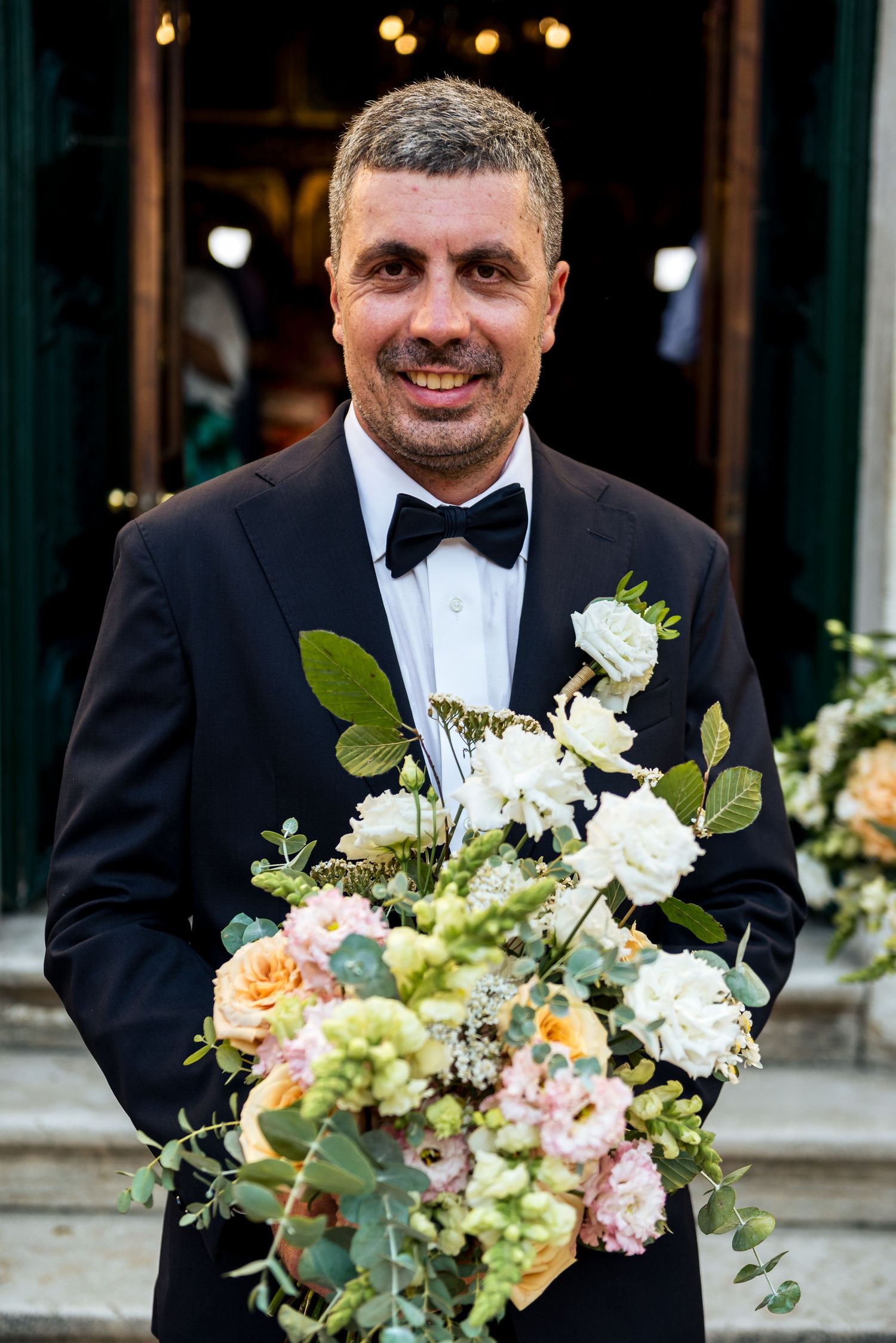 Groomsmen in black tuxedos hold beautiful pastel floral bouquets during an elegant wedding ceremony.