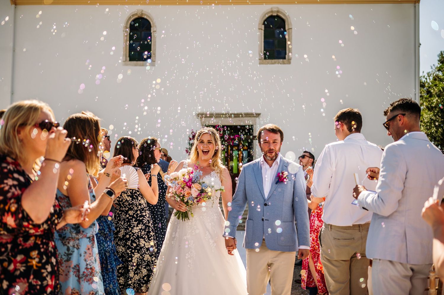Wedding celebration with guests throwing confetti outside a white Mediterranean church building.