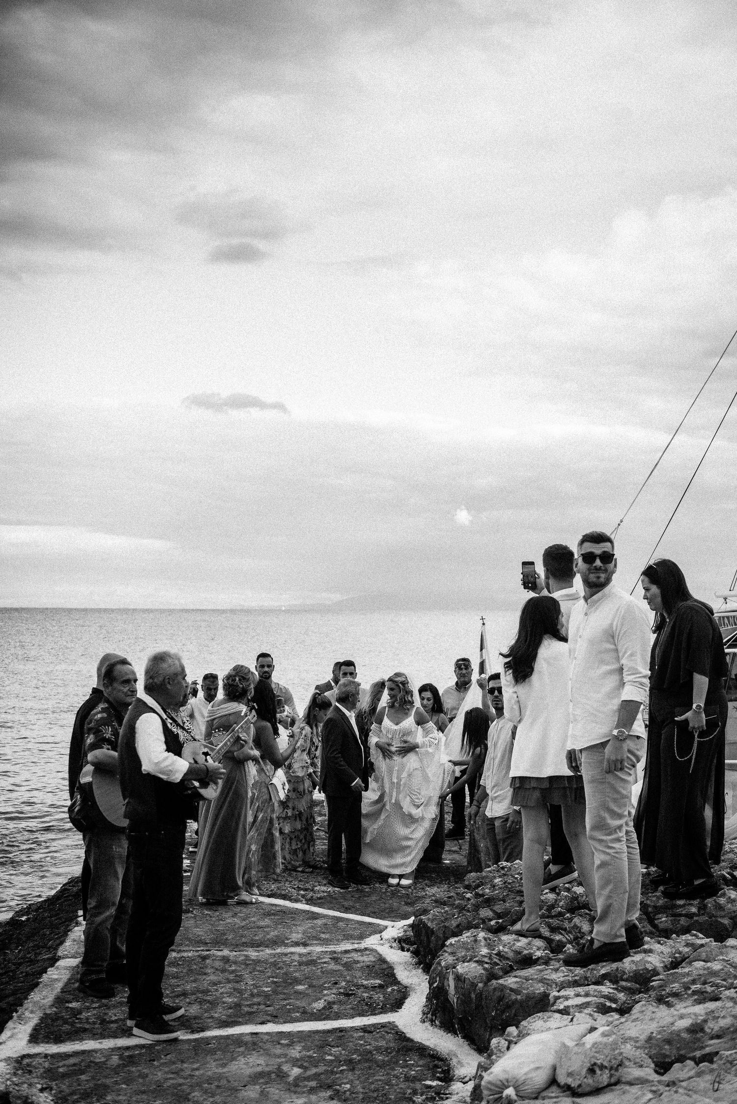 Wedding guests gather on a rocky coastal shoreline under cloudy skies for an outdoor ceremony.